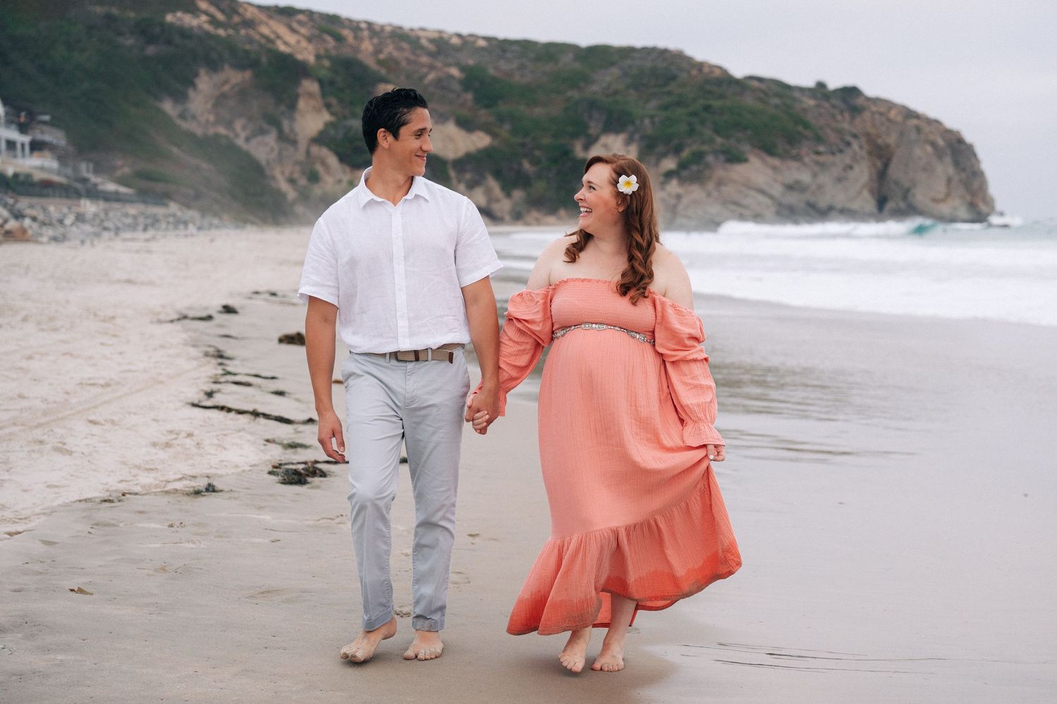 Couple walking hand in hand on a beach with coastal cliffs, wearing coordinated white and coral outfits.