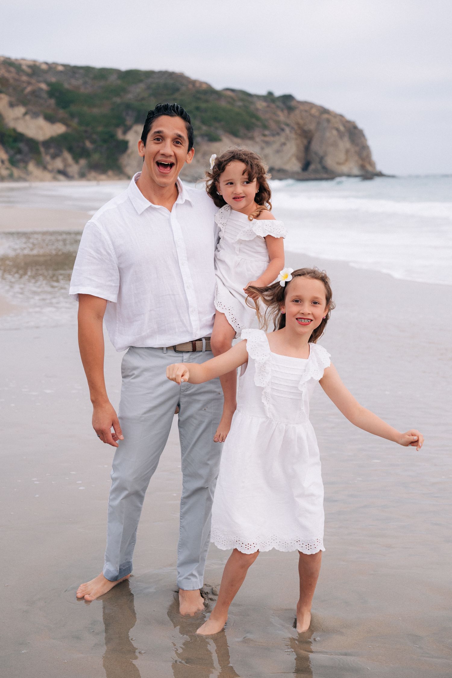 Family wearing coordinated white outfits smiles and poses together on a scenic beach with cliffs in the background.
