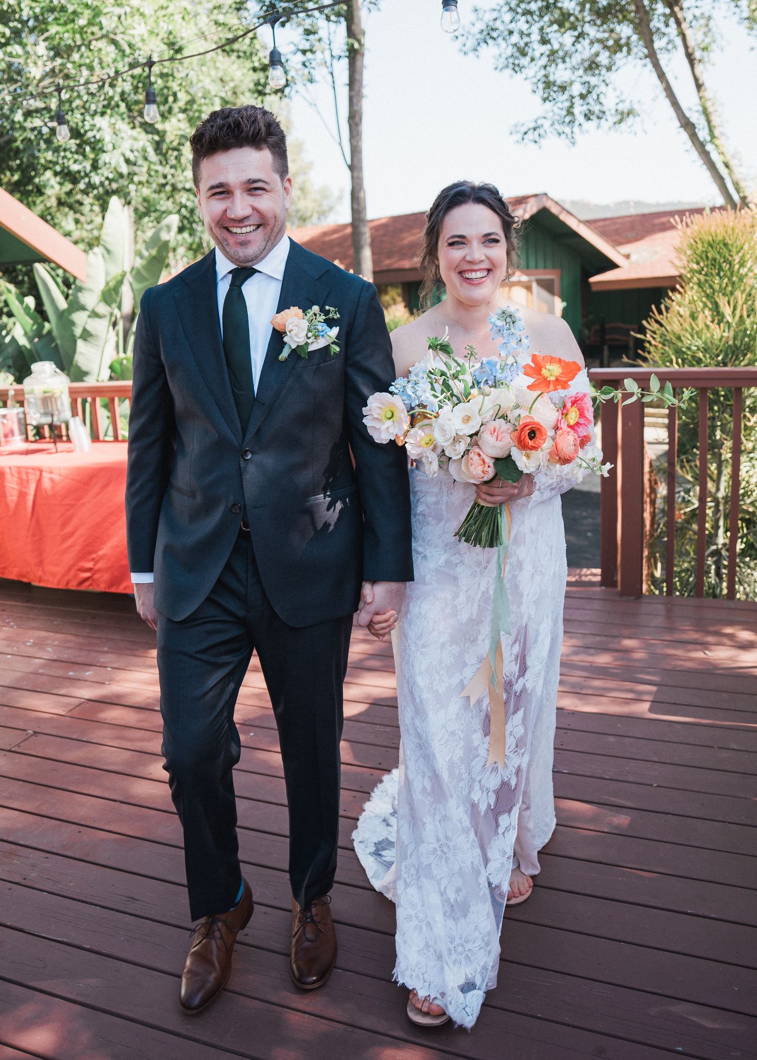 A wedding couple stands on a wooden deck with coral floral decorations and trees in the background.