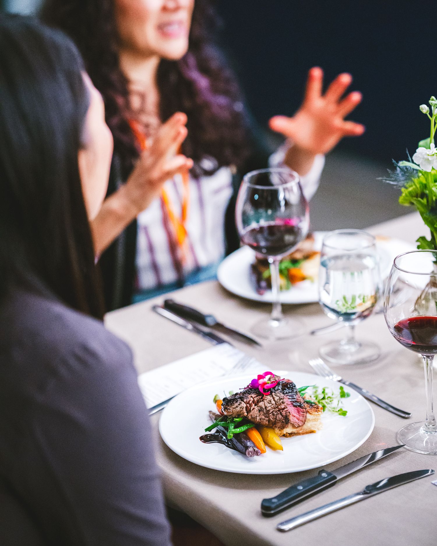Two diners engage in animated conversation over elegant plated dishes and wine glasses at an upscale restaurant table.