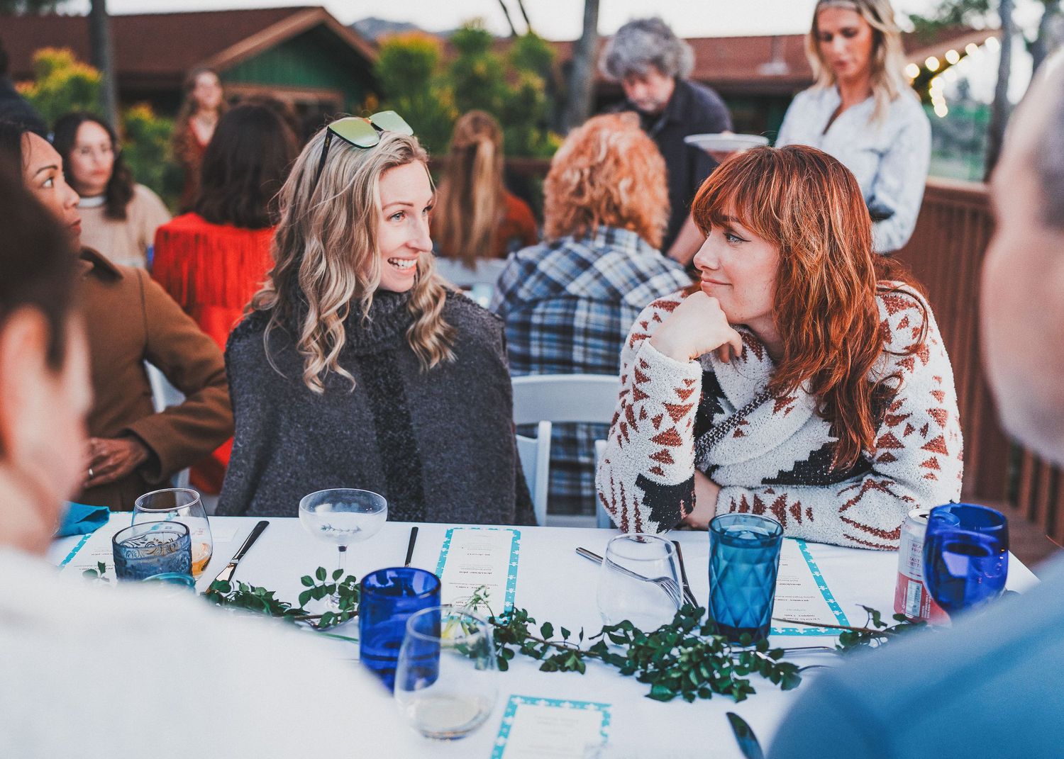 Friends engage in animated conversation at an outdoor table decorated with greenery and blue glassware during a gathering.