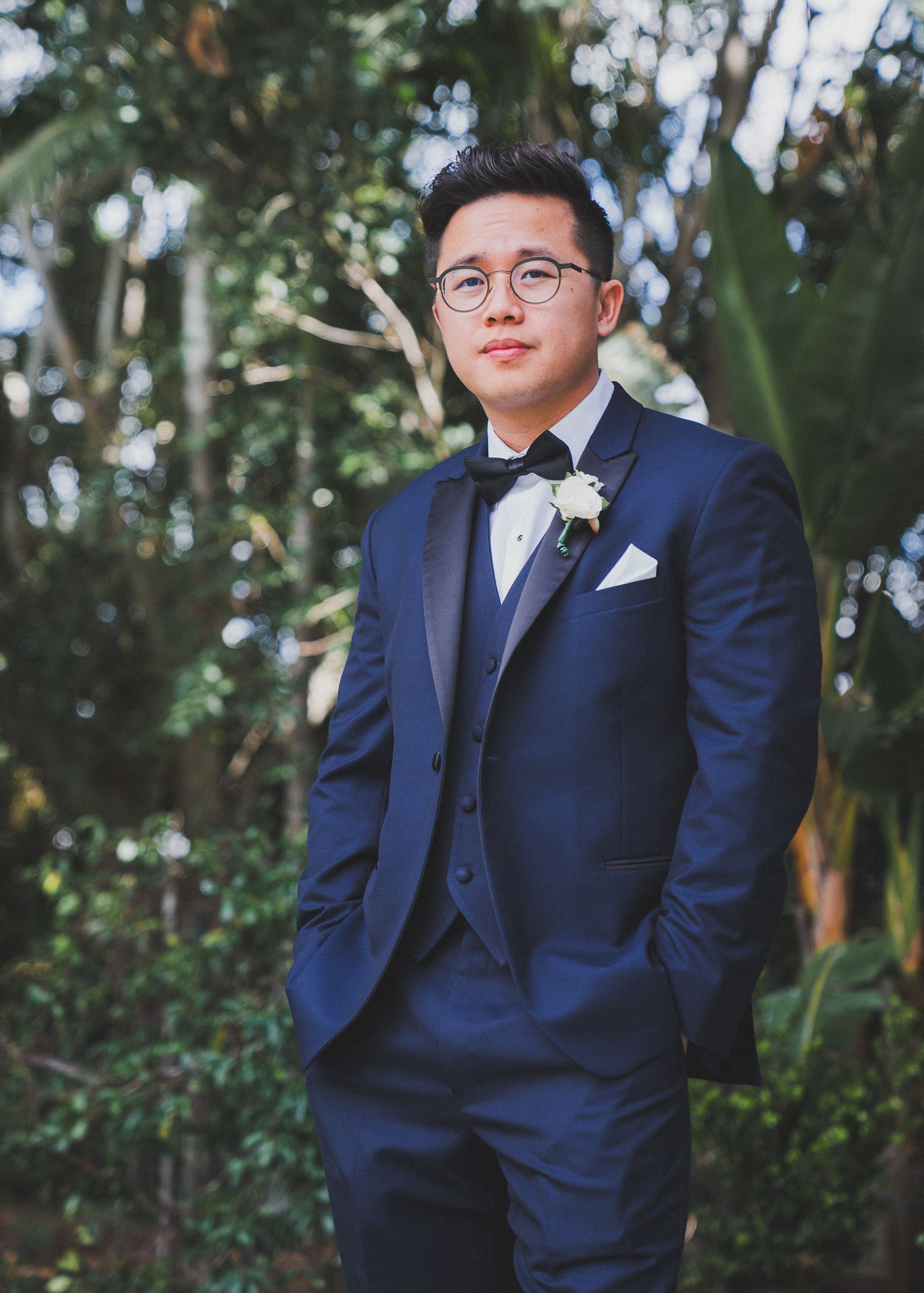 A smartly dressed person in a navy blue tuxedo with bow tie and glasses poses outdoors against lush greenery.