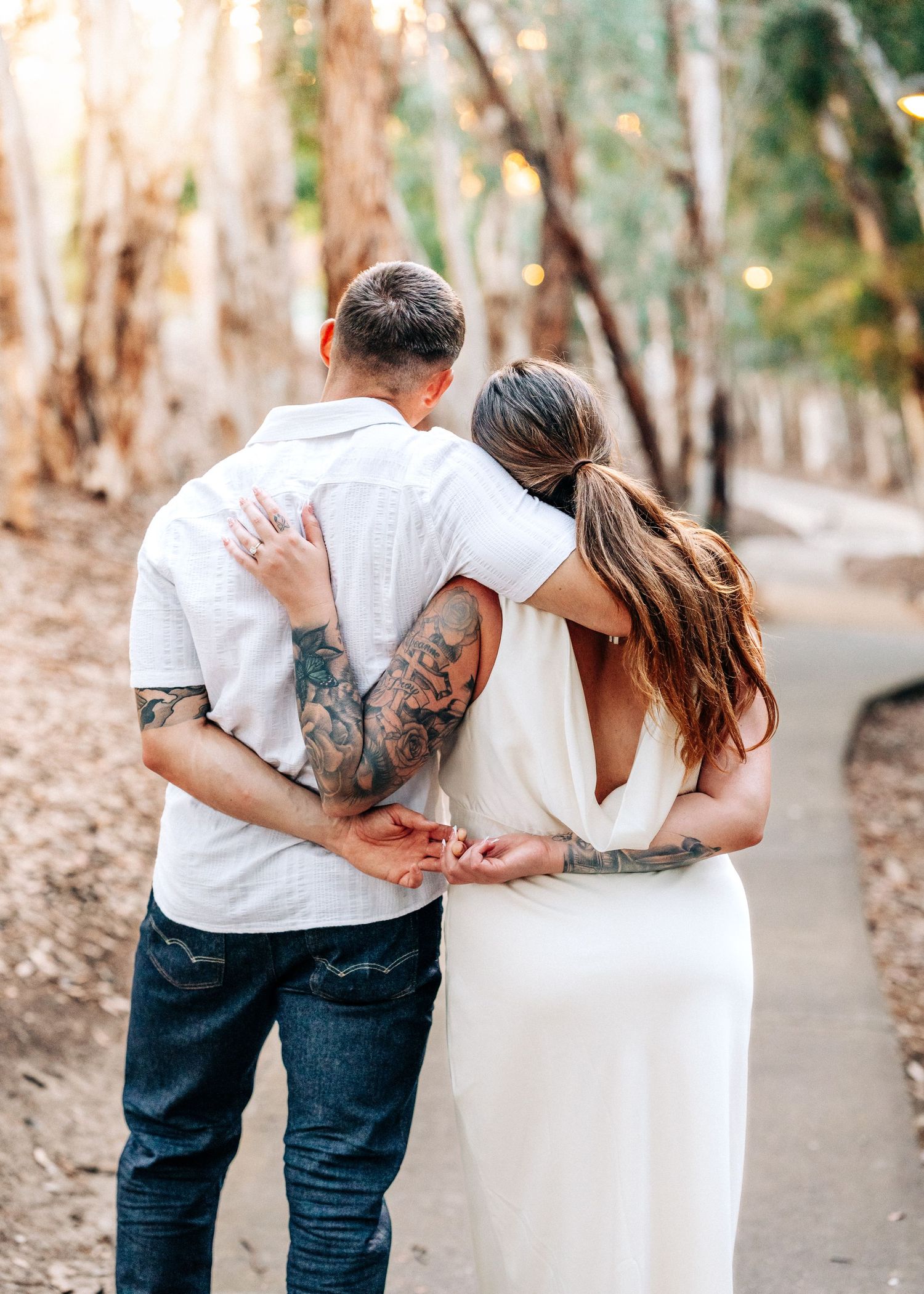 A couple embraces while walking along a tree-lined path with gum trees and fallen leaves.