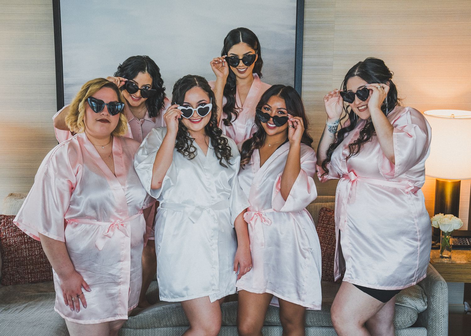 Wedding party wearing matching white outfits and sunglasses pose together against wall at sunset.