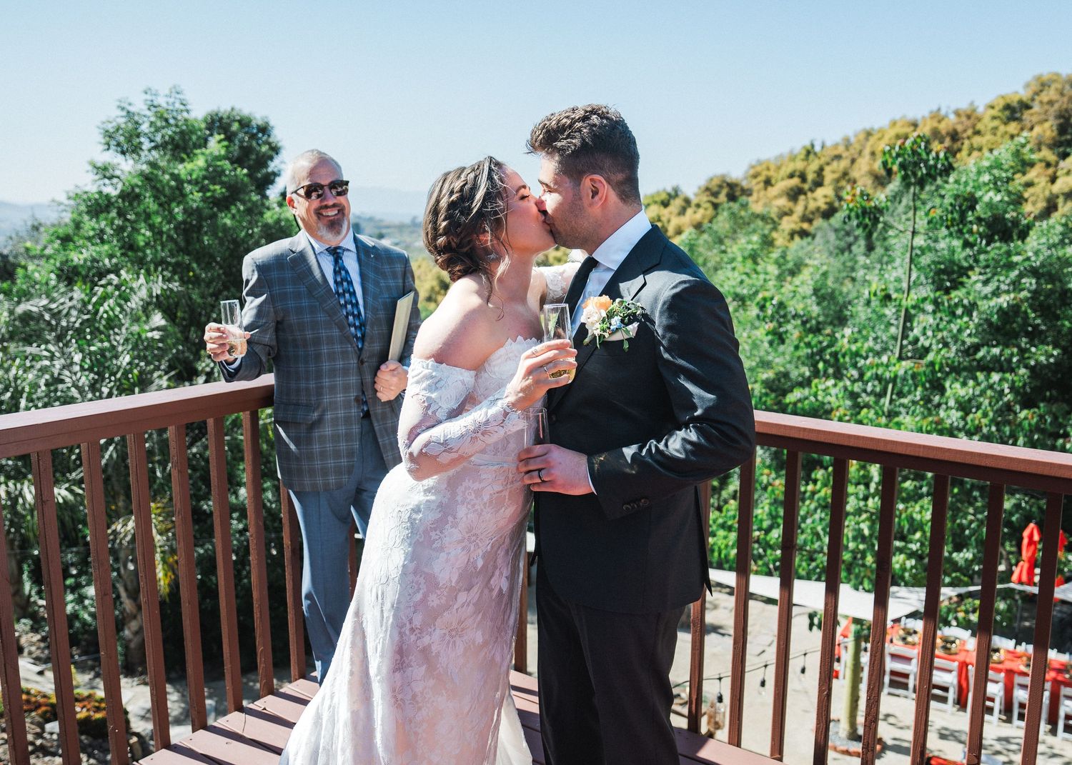 An outdoor wedding ceremony takes place on a wooden deck surrounded by lush greenery.