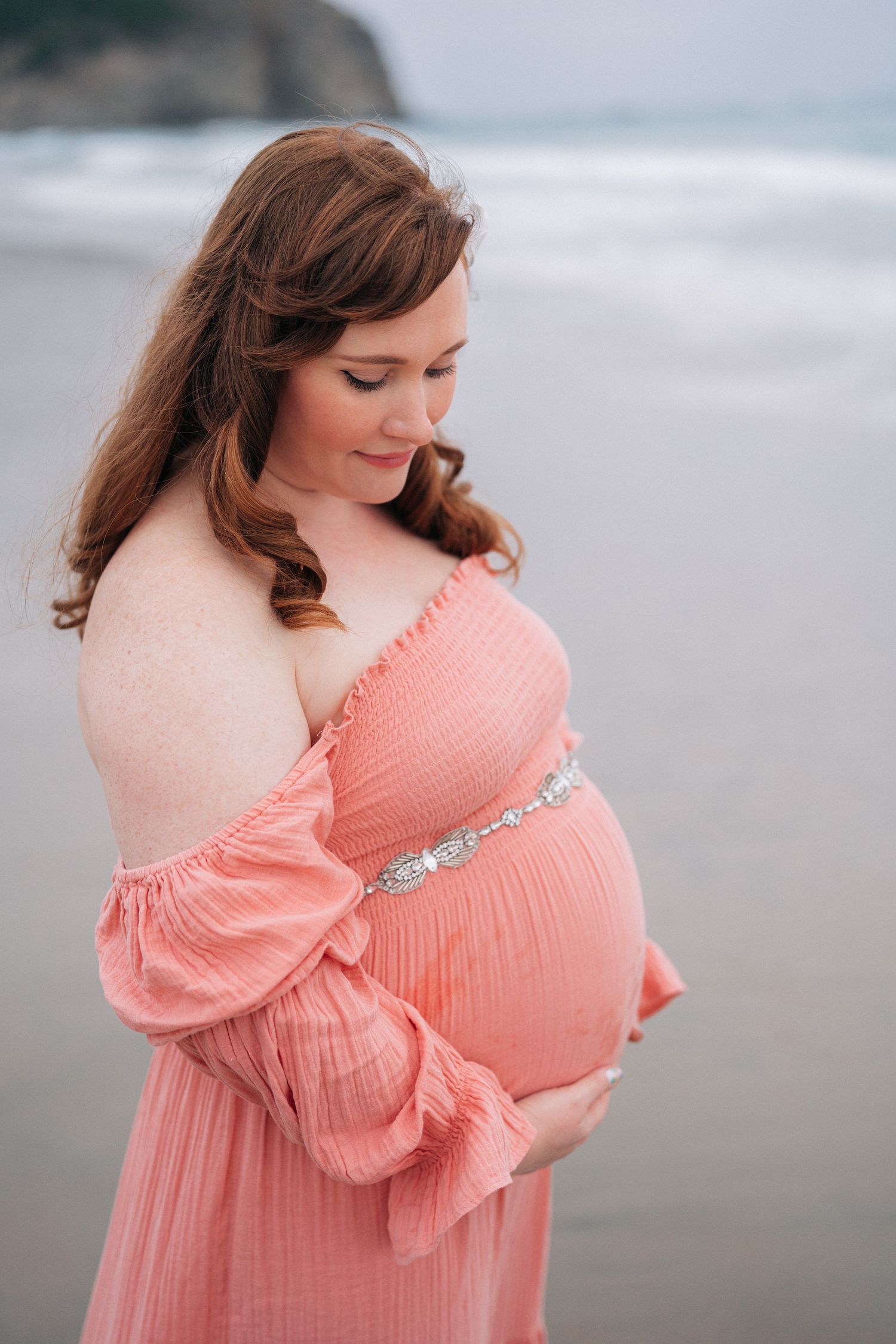 An expectant mother in a flowing coral dress poses on the beach with coastal cliffs in the background.