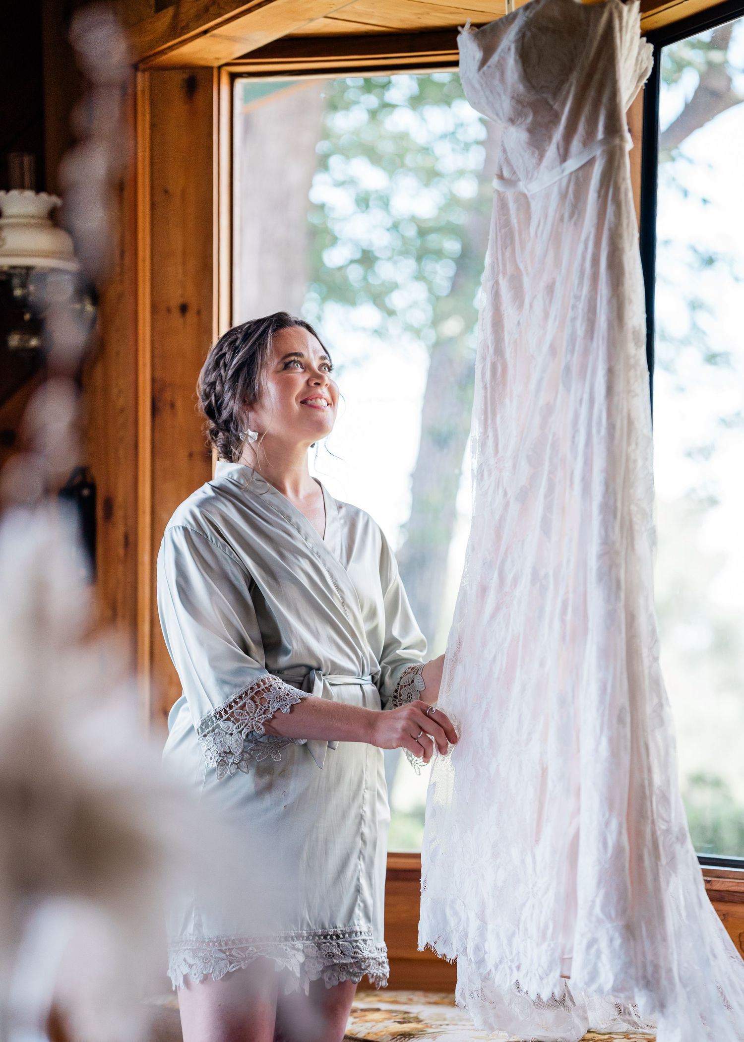 A white lace wedding dress hangs by a window as morning light streams in.