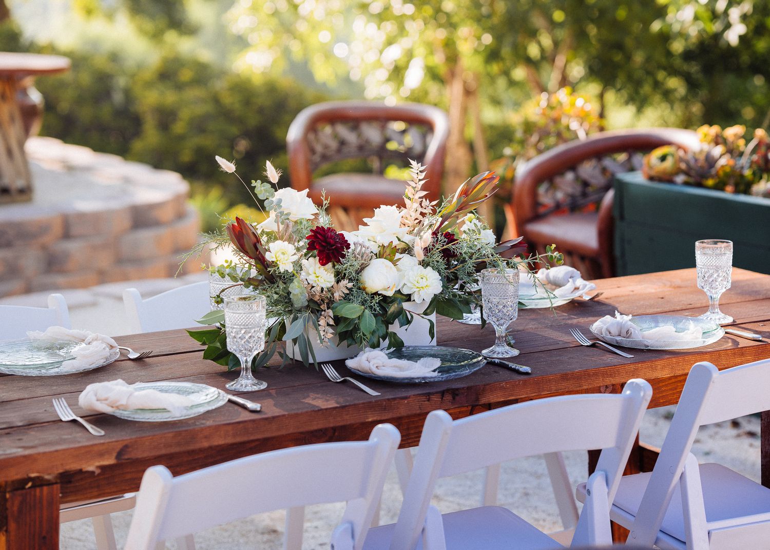 Elegant outdoor table setting with rustic wooden table, white chairs and floral centerpiece at garden wedding reception.