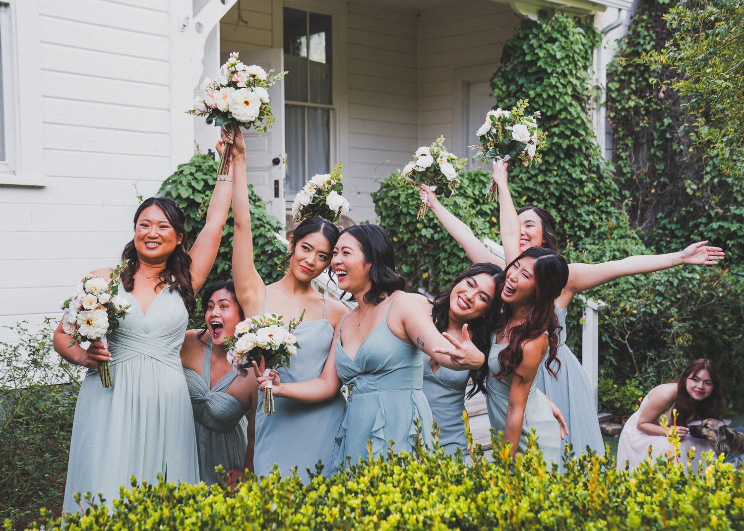 Bridesmaids in light blue dresses celebrating joyfully in front of white building.
