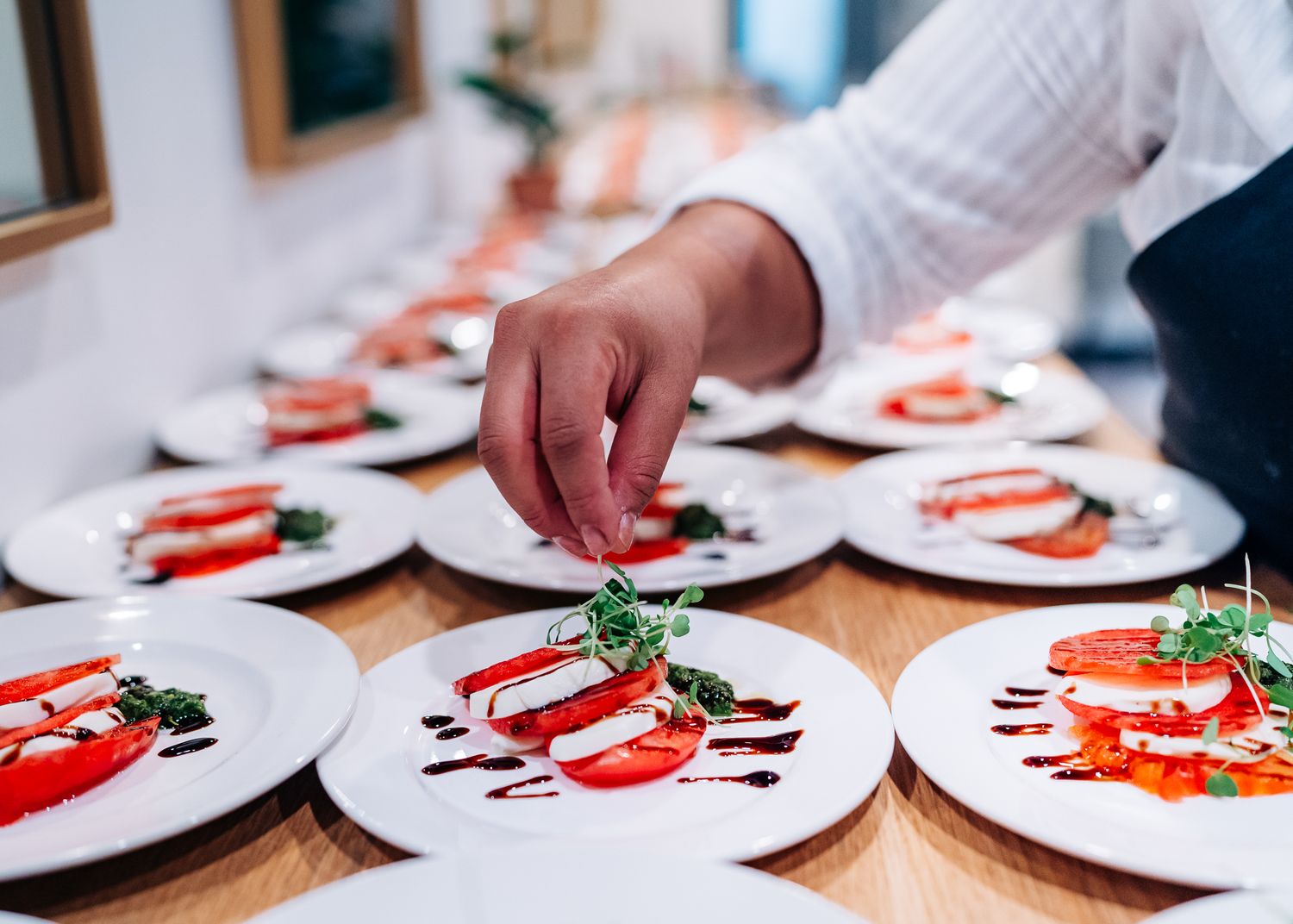 Multiple white plates of caprese salad with tomatoes and balsamic reduction being prepared in a restaurant.