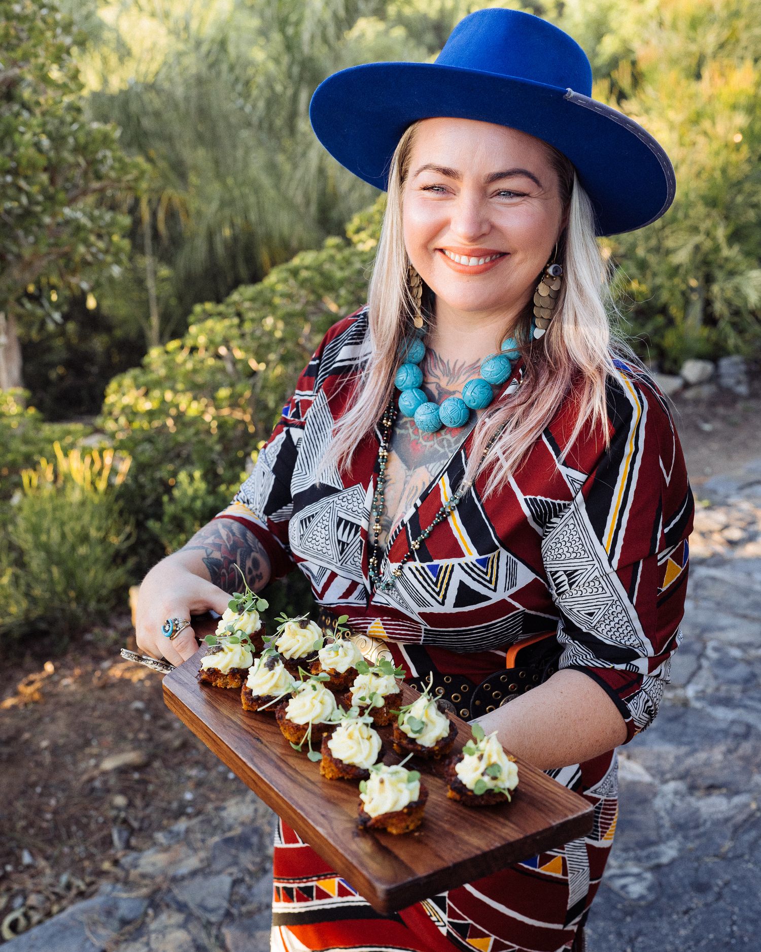 A person wearing a bright blue hat and turquoise jewelry holds a wooden tray of freshly baked appetizers in an outdoor setting.