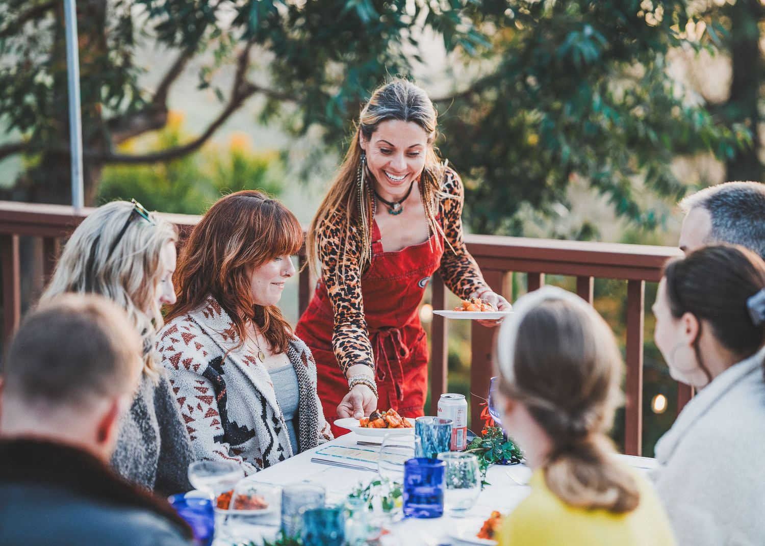 A gathering of people sharing a meal outdoors on a wooden deck with table settings and glassware.