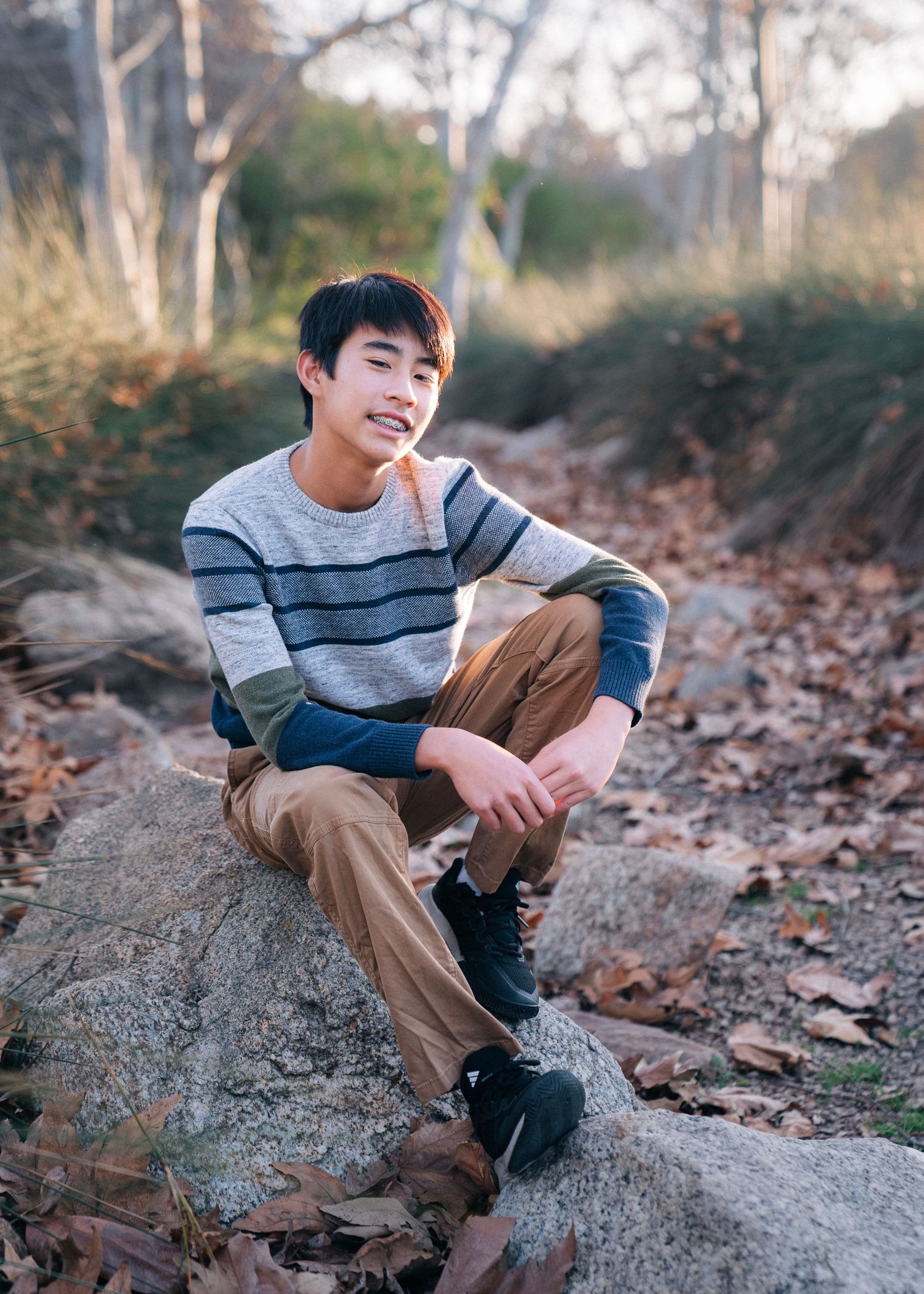 A person in a striped sweater sits casually on a rock among fallen leaves during autumn.