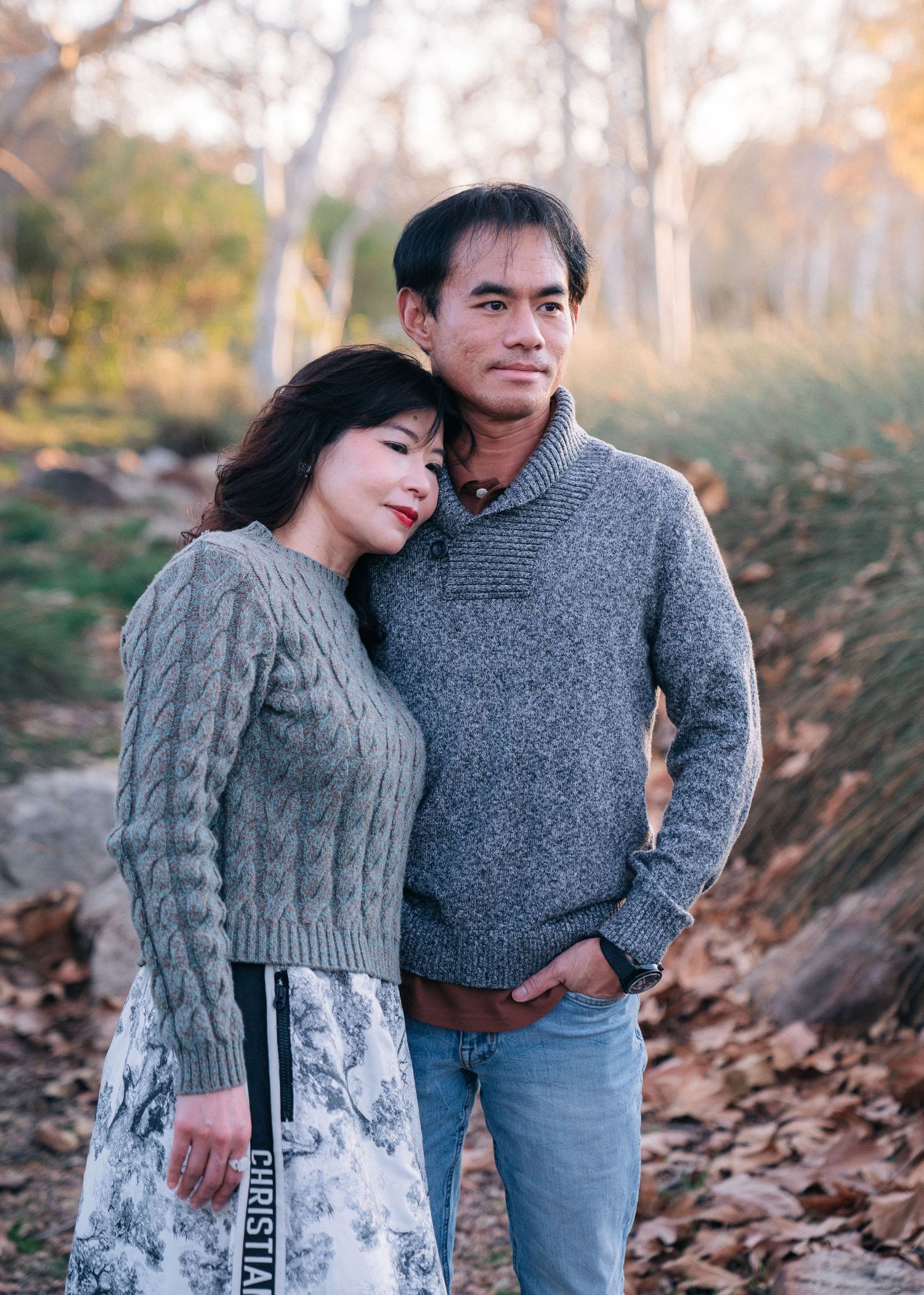 A couple in cozy sweaters share an intimate moment outdoors during autumn, surrounded by trees and fallen leaves.
