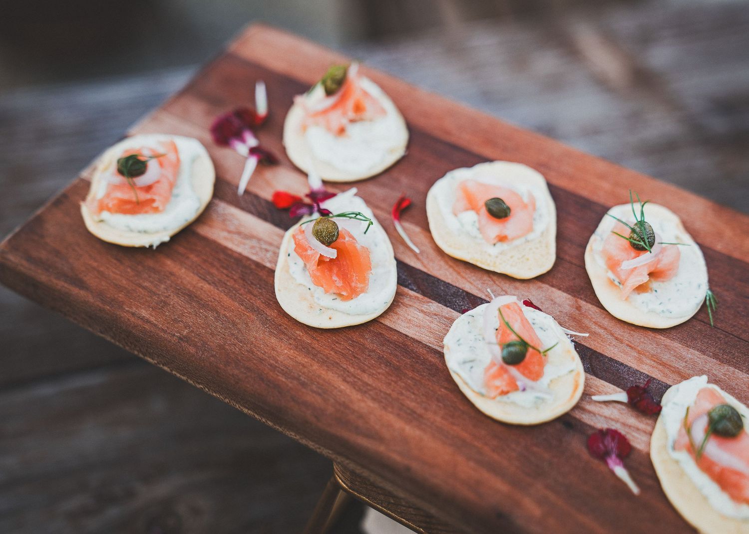 Small smoked salmon and cream cheese appetizers arranged on a wooden serving board with capers and fresh herbs.