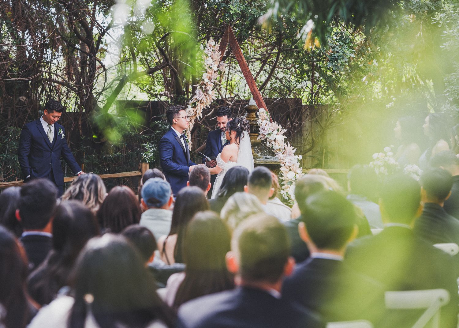 Wedding ceremony guests seated outdoors under trees with natural sunlight filtering through green foliage.
