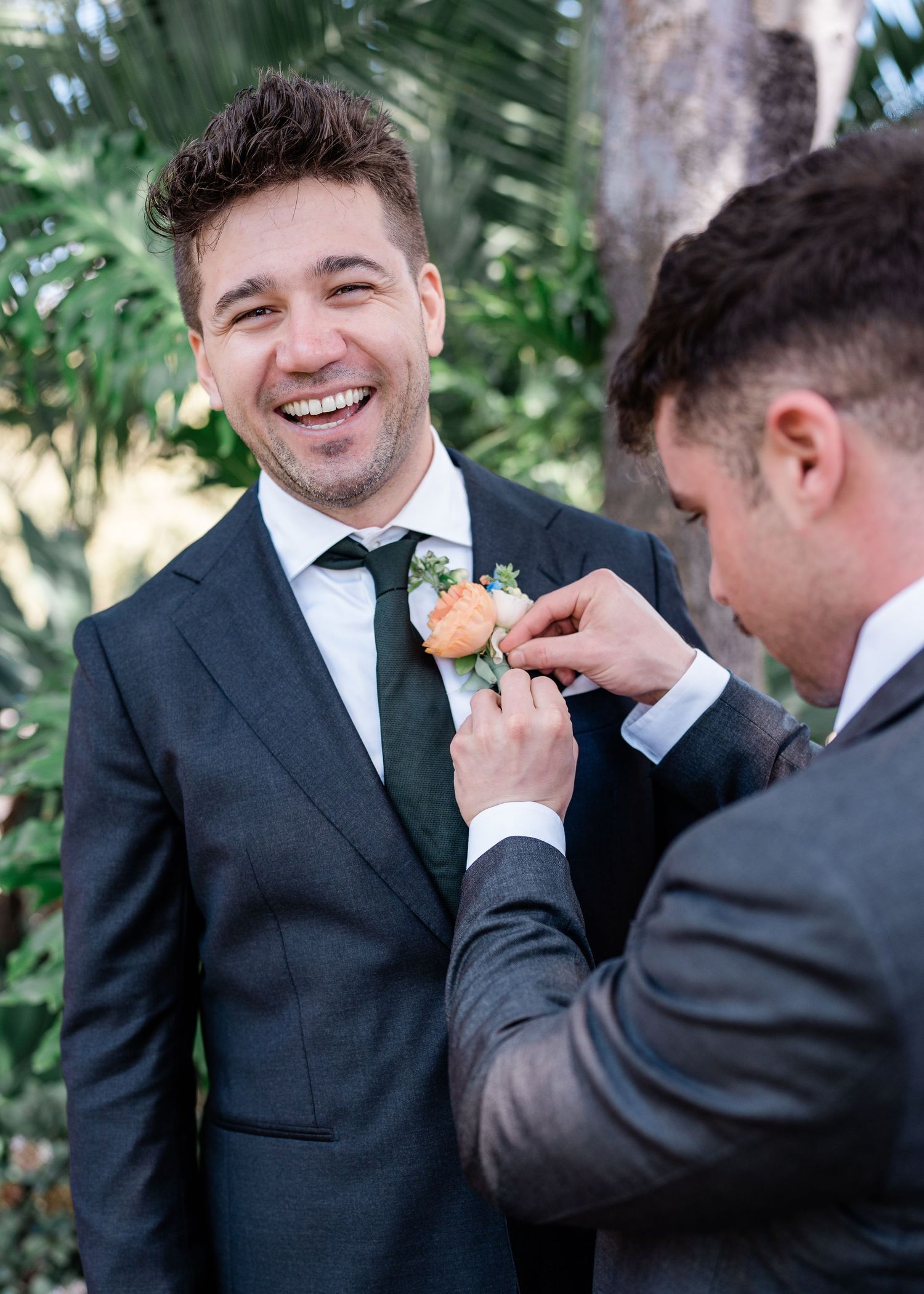 A groomsman helps adjust a coral boutonniere on the groom's lapel during an outdoor wedding ceremony.