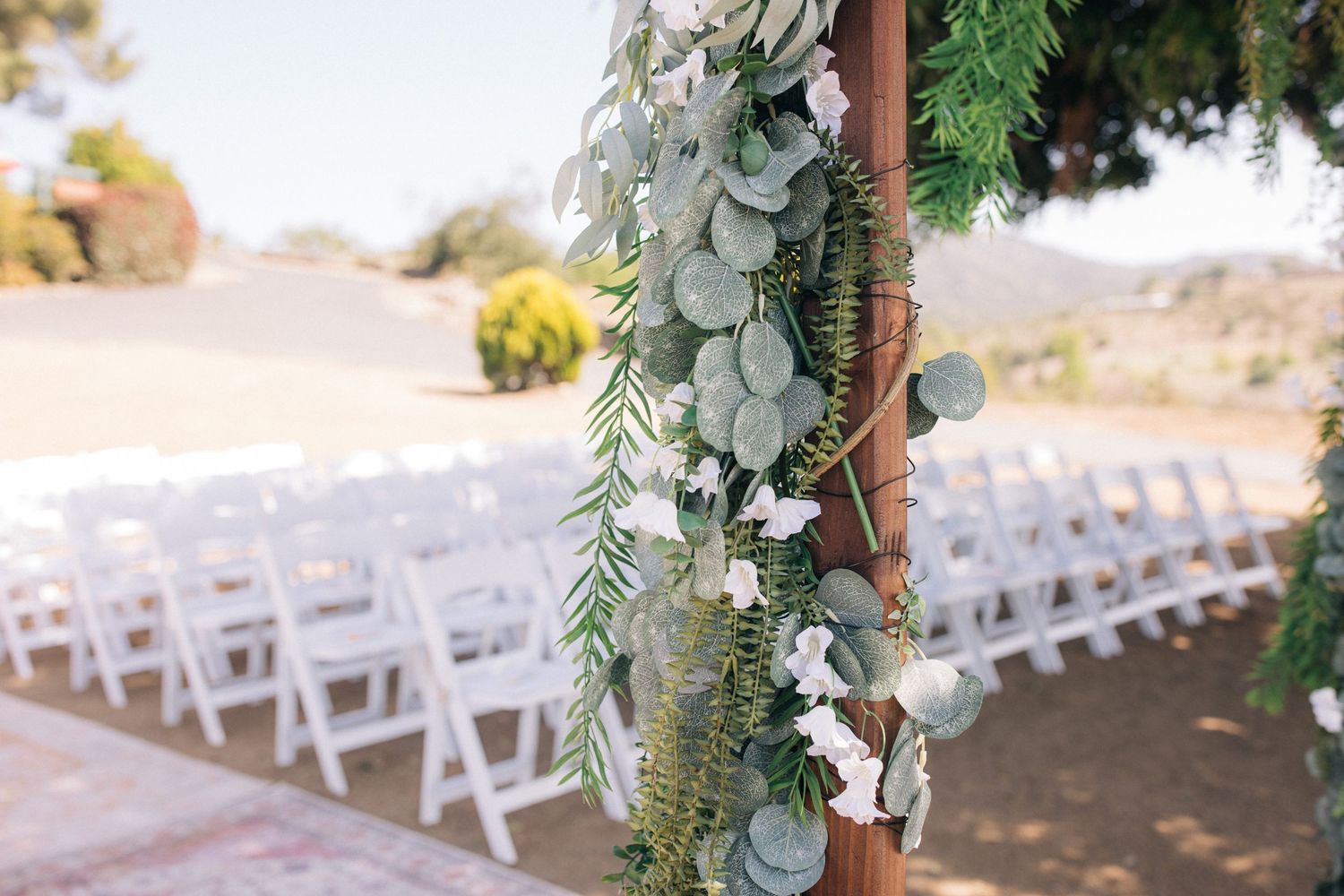Outdoor wedding ceremony setup with white chairs and greenery arch decorations against a rural landscape.