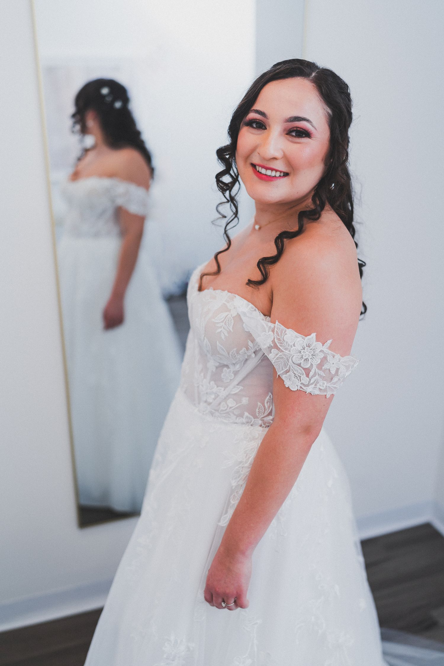 A bride in an off-shoulder white lace wedding dress with curled hair smiles while her reflection shows in a mirror behind her.