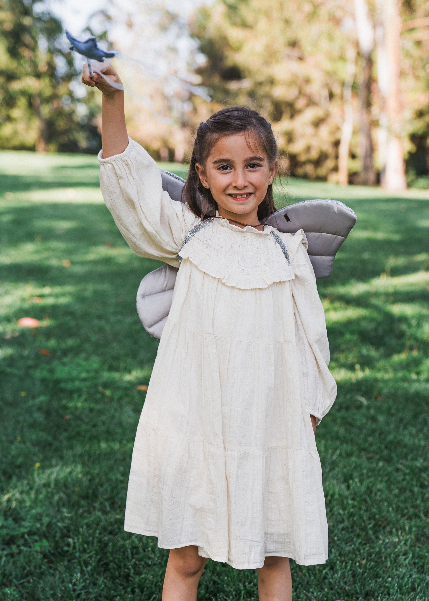 Child in cream-colored prairie dress holds arm up while standing in a sunny green field on a bright day.