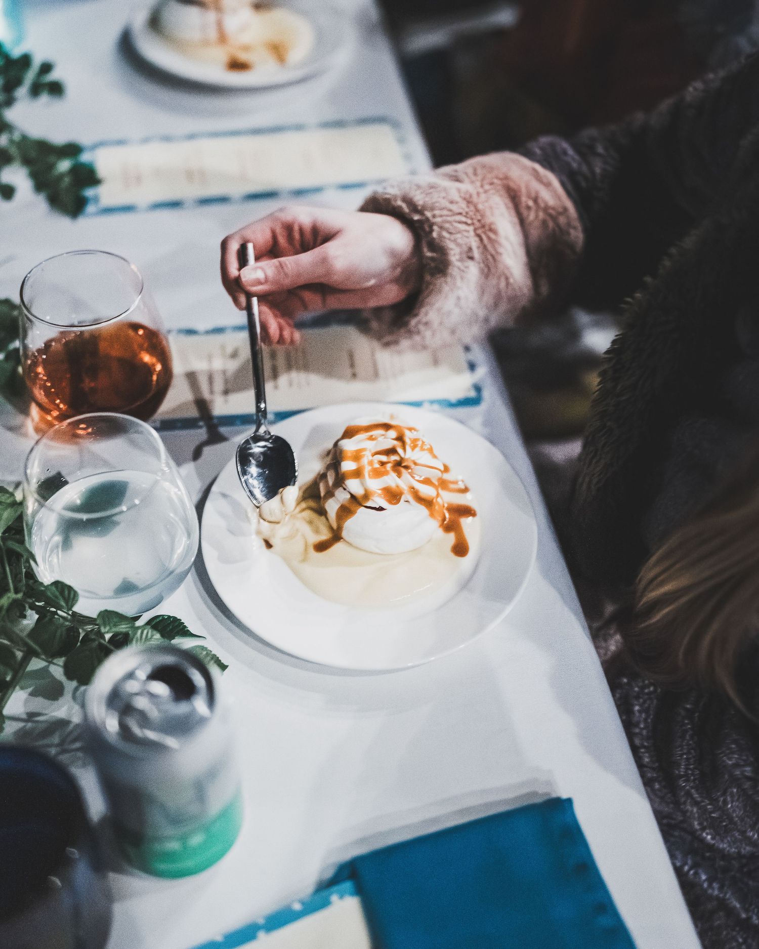 Hand drizzling caramel sauce over vanilla ice cream dessert with wine and water glasses on white table setting.