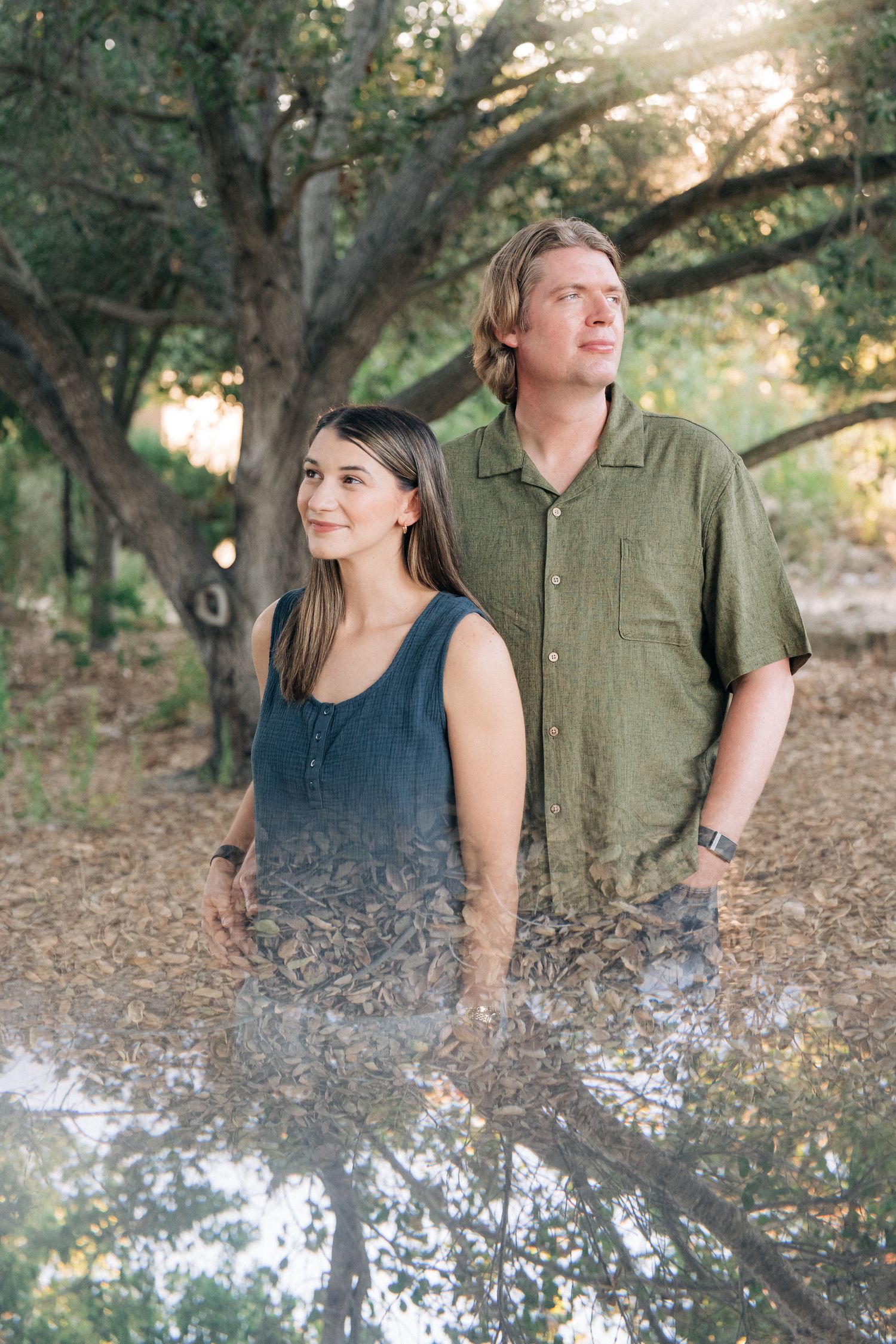 A couple stands together outdoors on a wooded trail during golden hour with a dreamy, flare-lit natural backdrop.