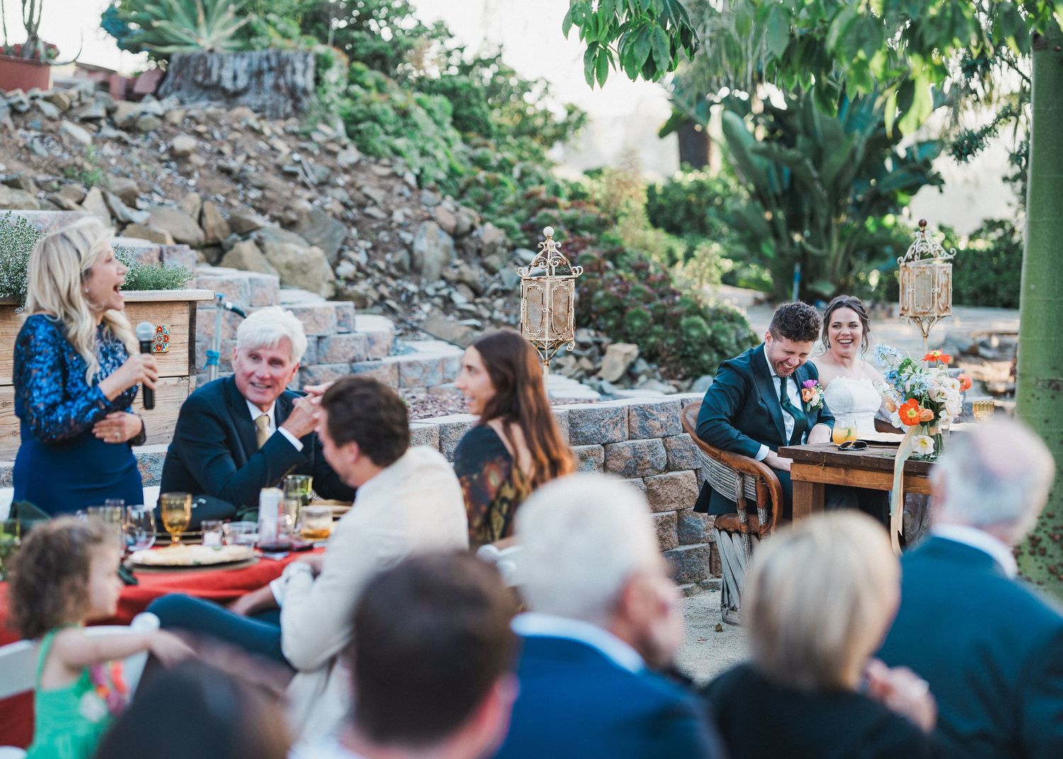 Outdoor wedding reception with guests seated under trees and stone wall backdrop.