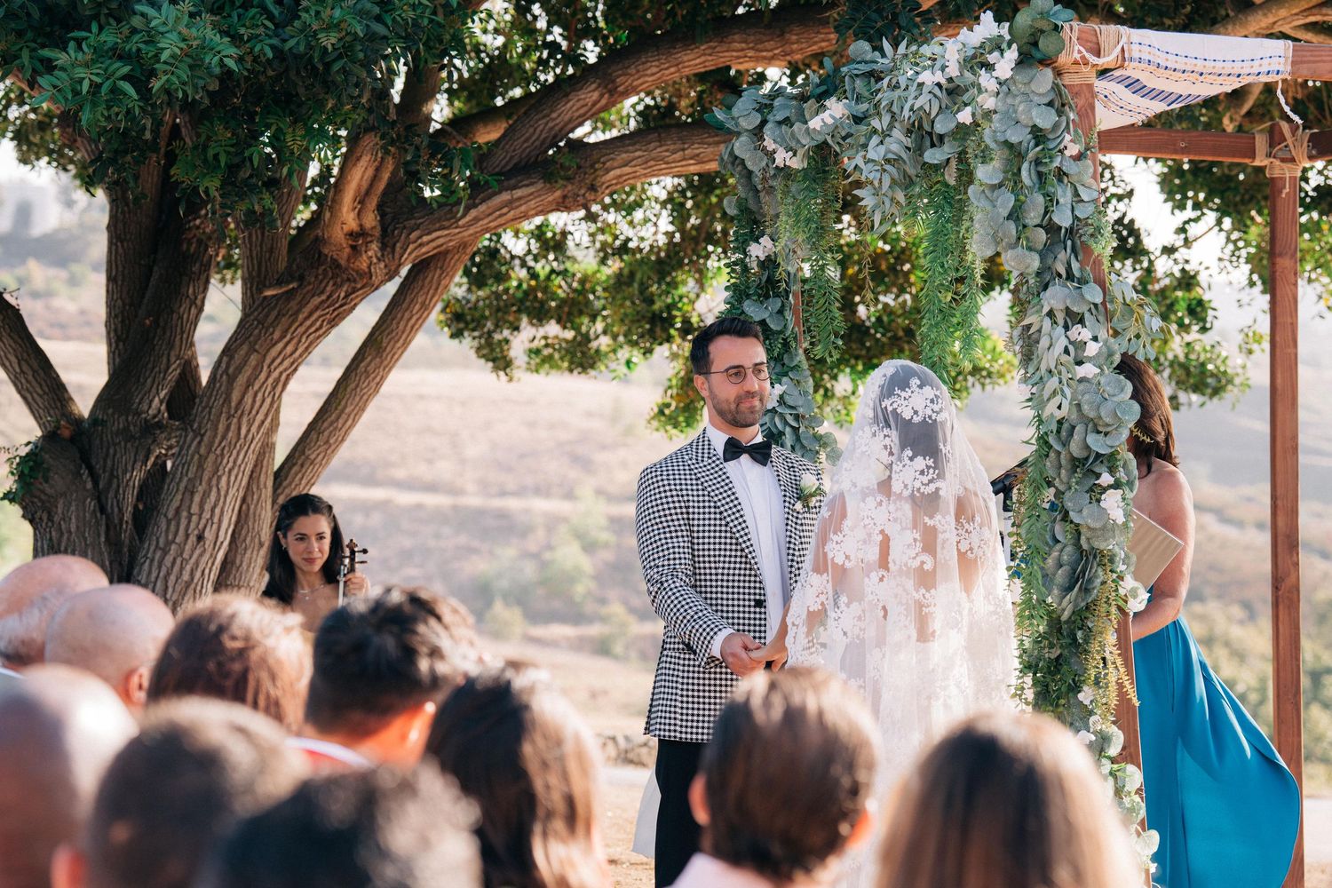 A romantic outdoor wedding ceremony under a tree with floral decorations and gathered guests.