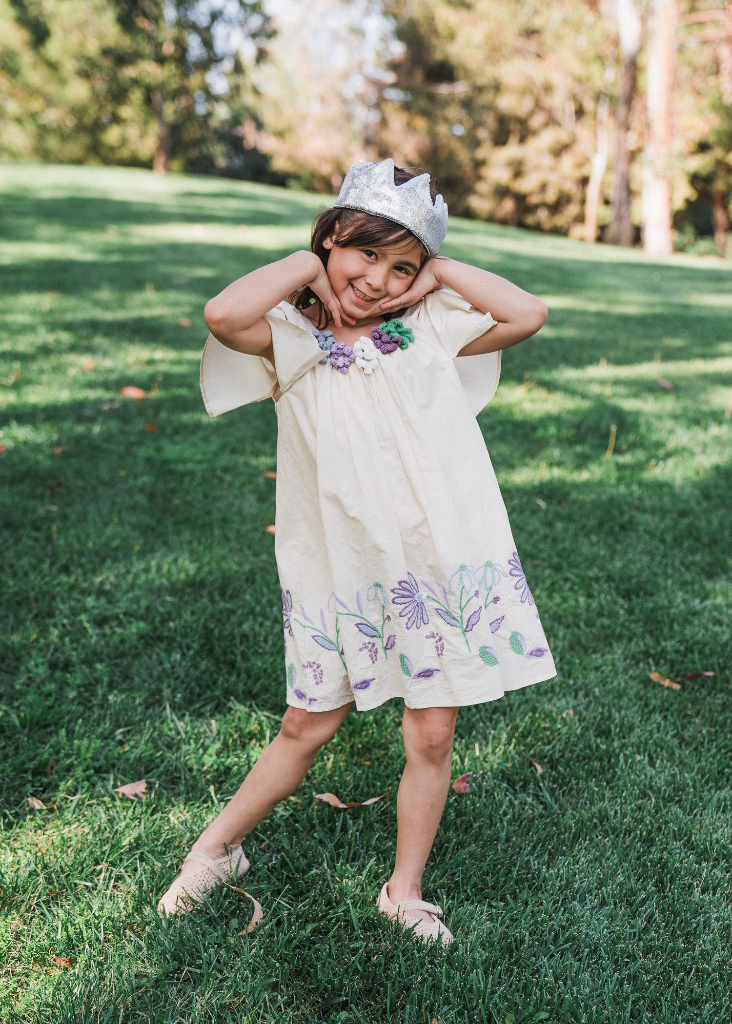 A child in a white dress with purple floral embroidery poses playfully on green grass in a sunny park setting.