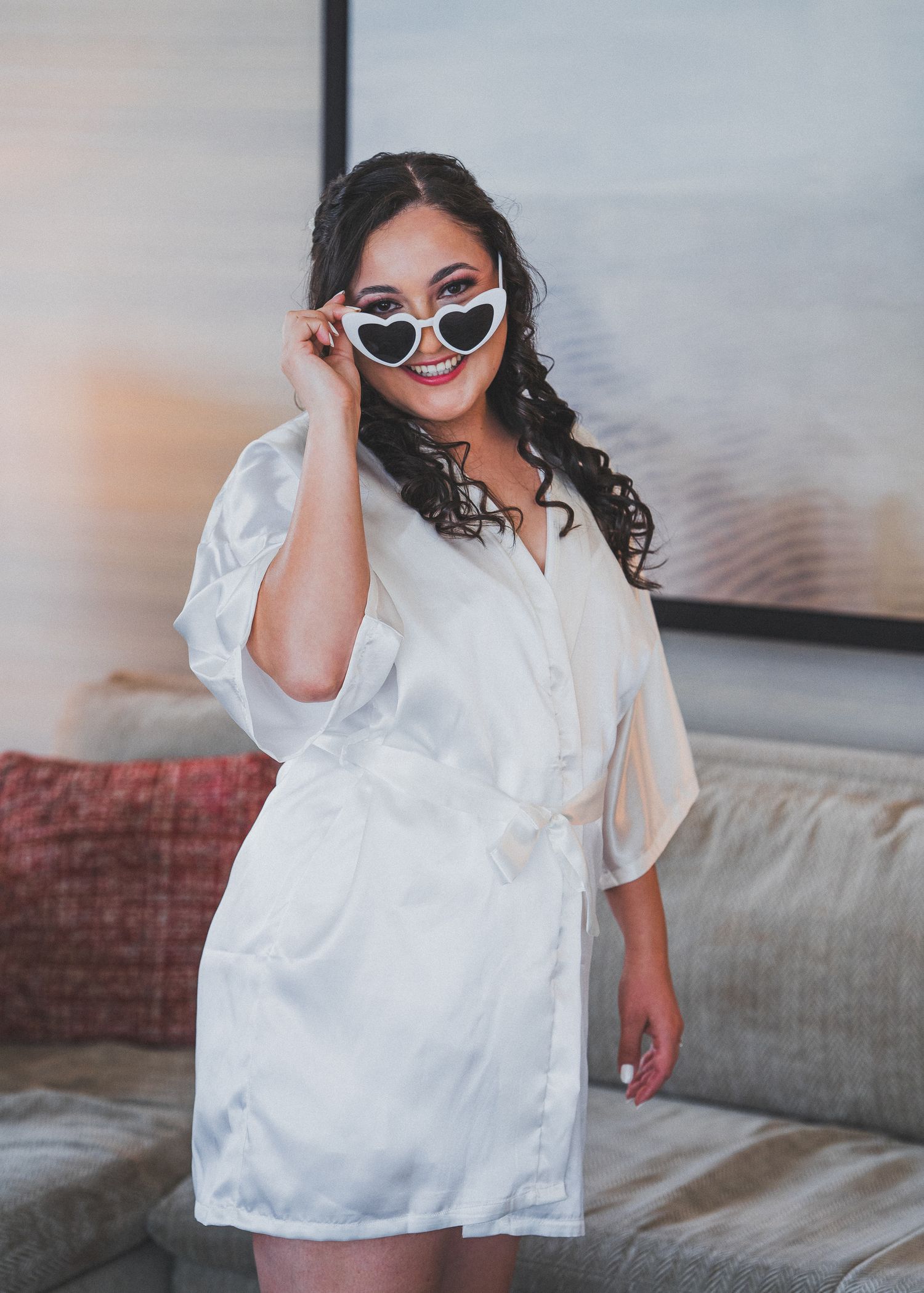 Fashion model on gray couch wearing stylish white button down shirt and dark sunglasses in lifestyle setting.
