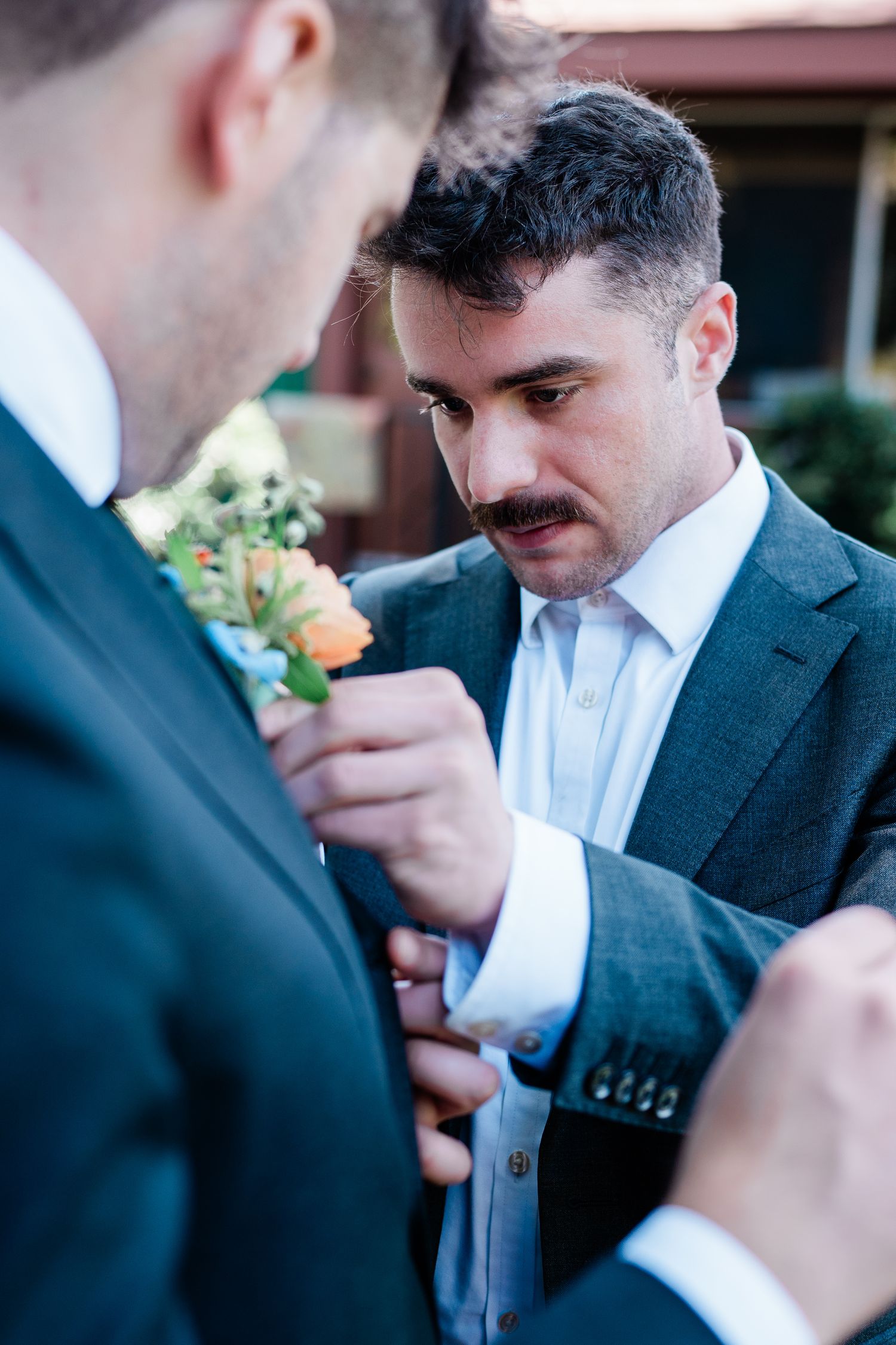 Wedding party members in dark suits help pin boutonniere flowers for formal event.