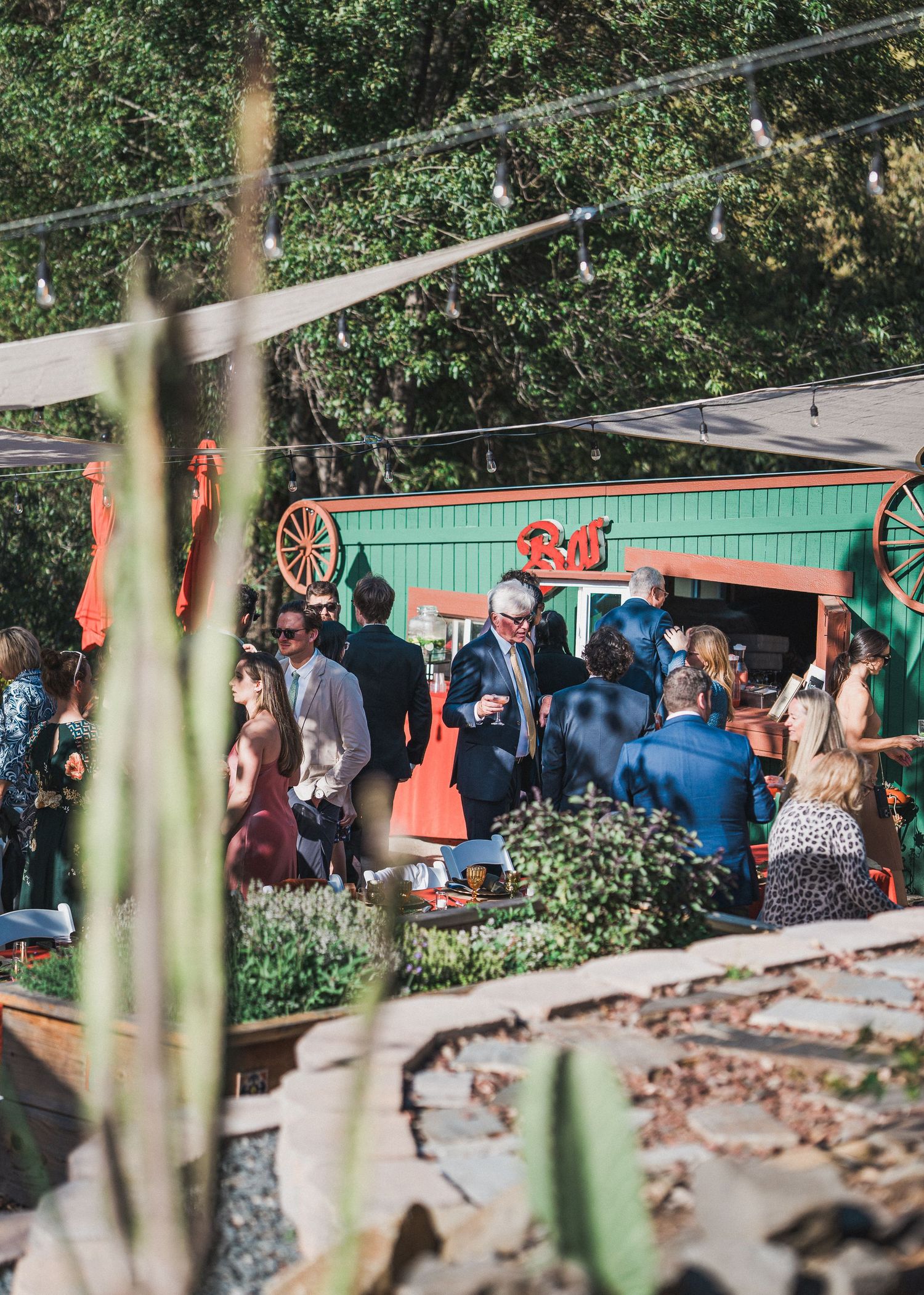 People gathered at an outdoor party venue decorated with red wagon wheel accents and garden greenery on a sunny day.
