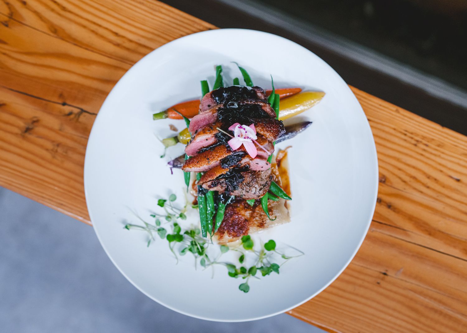 Artistically plated meat dish garnished with fresh herbs and colorful vegetables on a white plate against wooden surface.