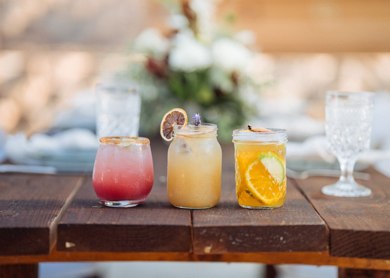 Three colorful summer cocktails in glass jars with citrus garnishes displayed on a rustic wooden table at an outdoor event.