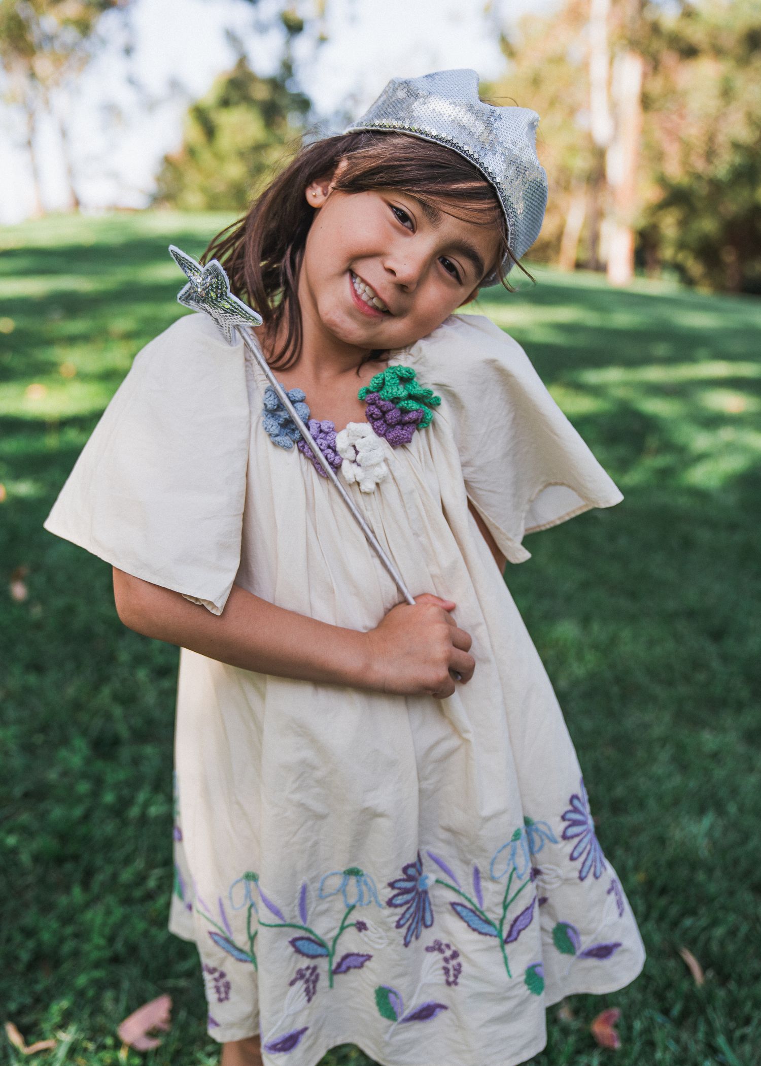 A child wearing a denim cap and white dress with purple floral embroidery poses playfully in a sunny garden setting.