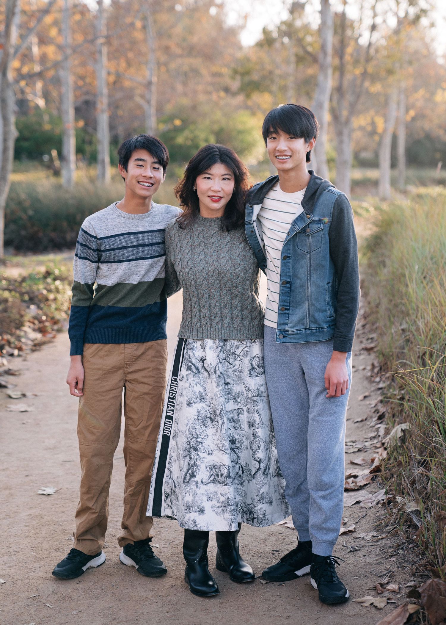 Three friends in casual attire stand together smiling on an autumn trail surrounded by fall foliage.