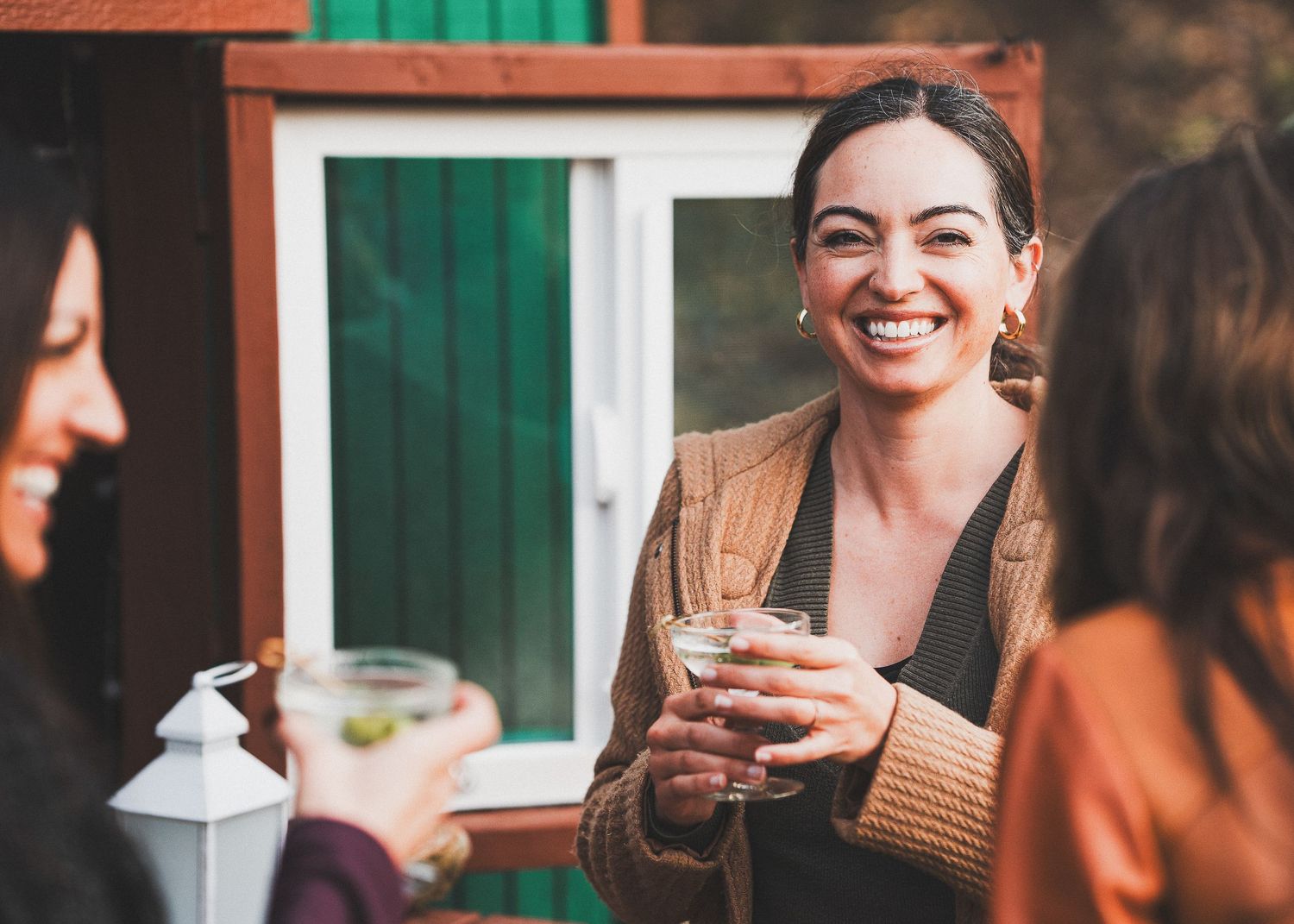 Three friends enjoying drinks and conversation while smiling and laughing together outdoors.
