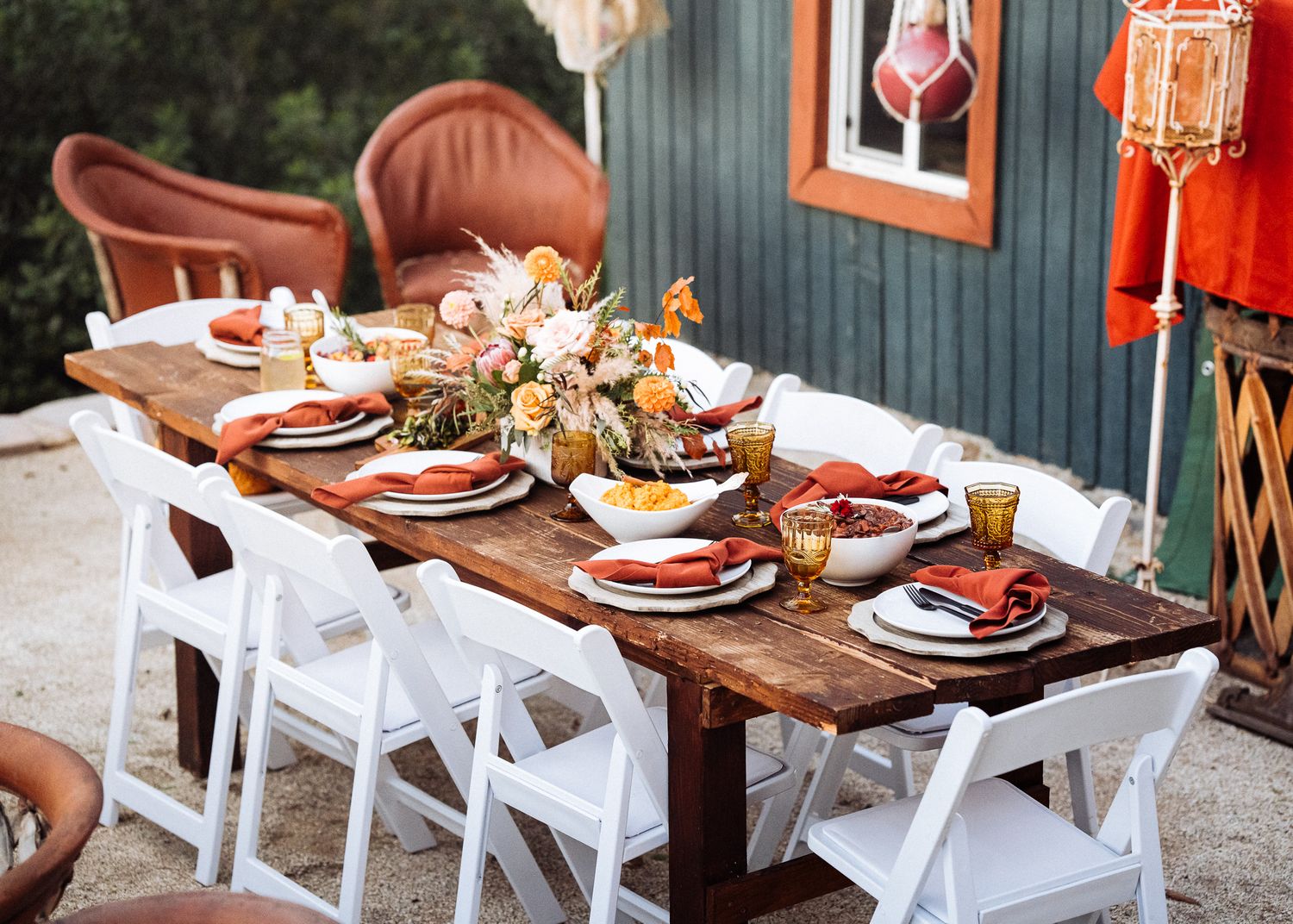 Rustic outdoor dining table with white chairs, floral centerpiece and place settings for a fall gathering.