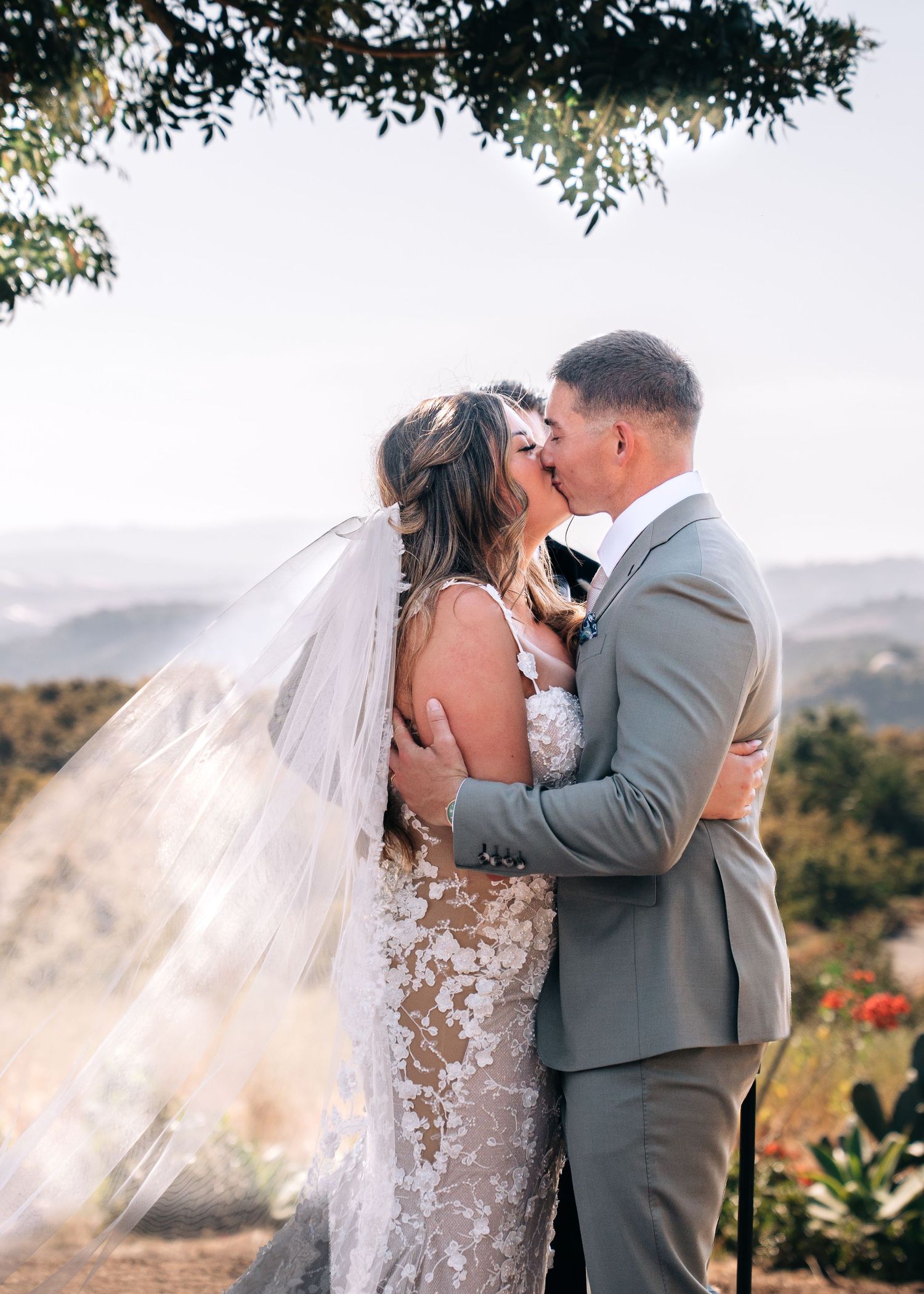 A romantic wedding photo showing a couple sharing a kiss outdoors with a scenic mountain backdrop.
