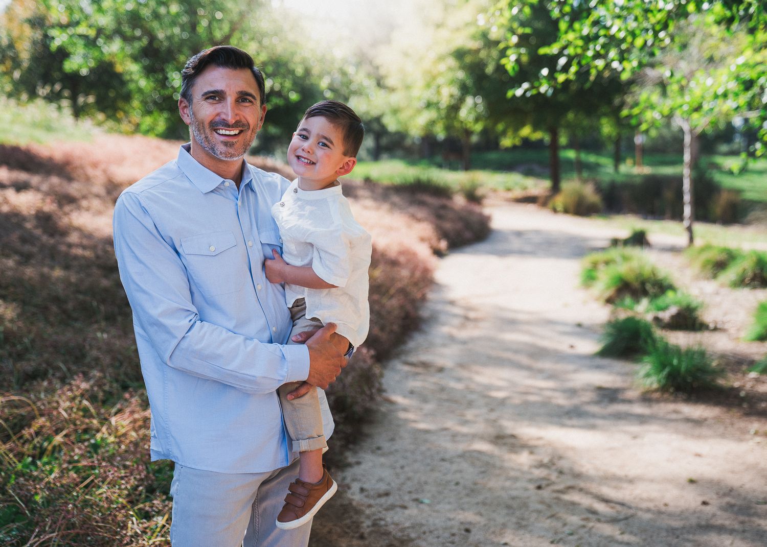 Two people share a warm embrace while standing on a sunlit dirt path in a lush garden setting.