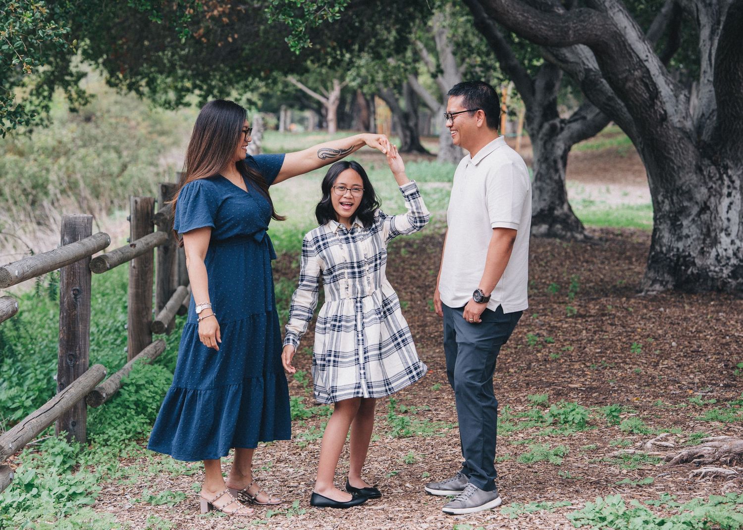 A family of three enjoys a candid moment together while standing near a wooden fence under shady trees in a park.