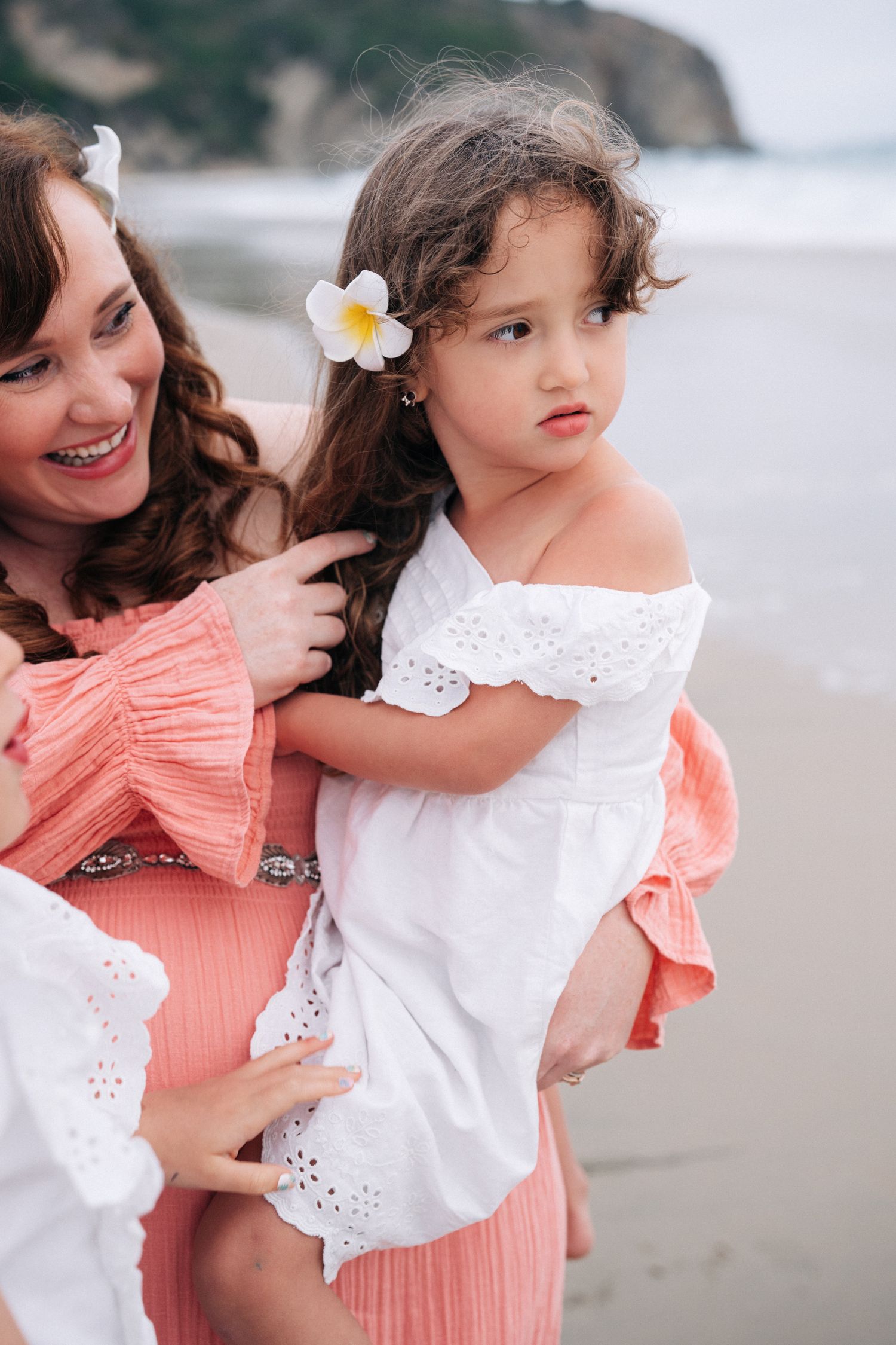 A mother and daughter share a tender moment on the beach with the little girl wearing a white dress and plumeria flower.