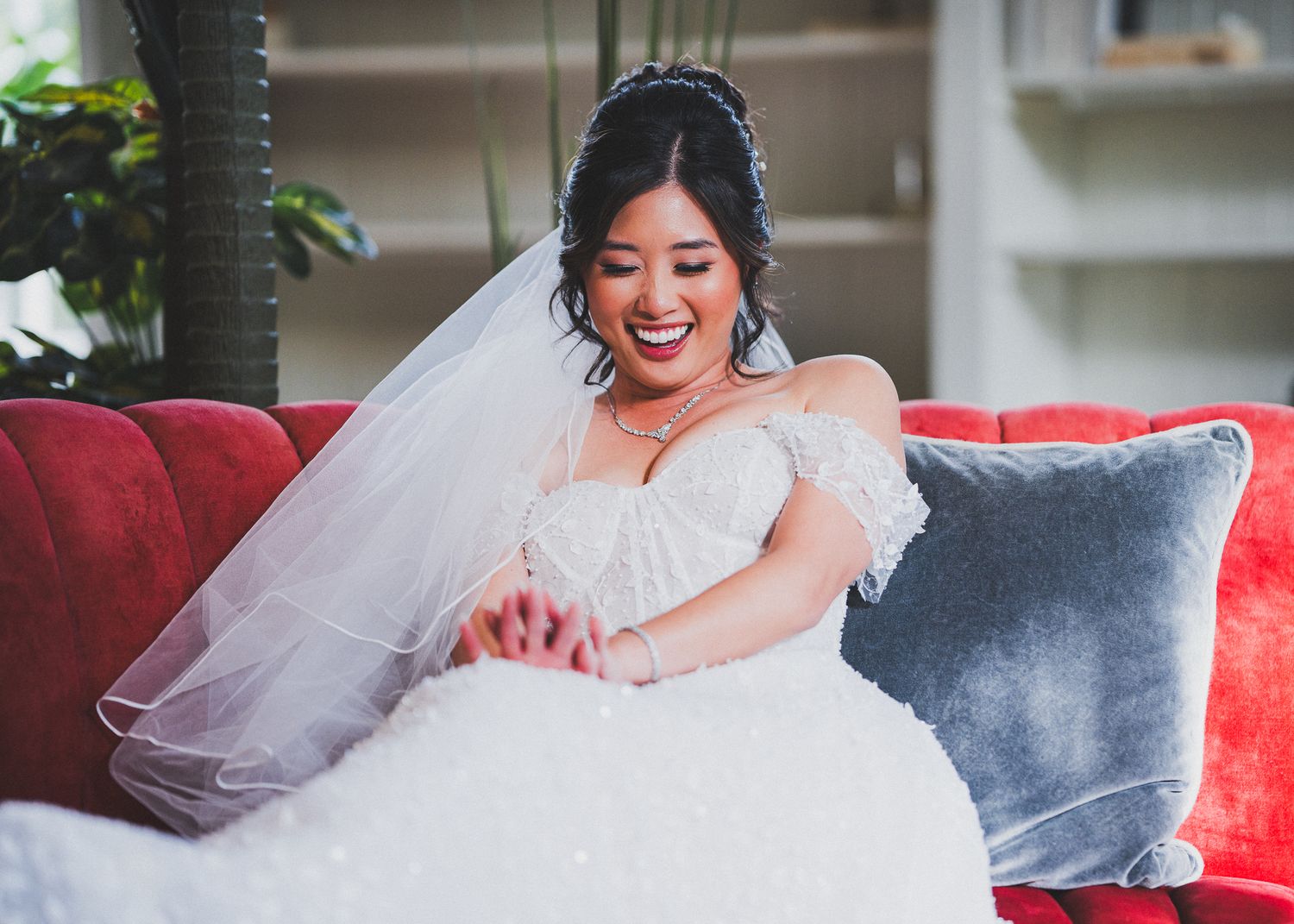 Close-up details of a bride's flowing white lace wedding gown on a red velvet sofa.