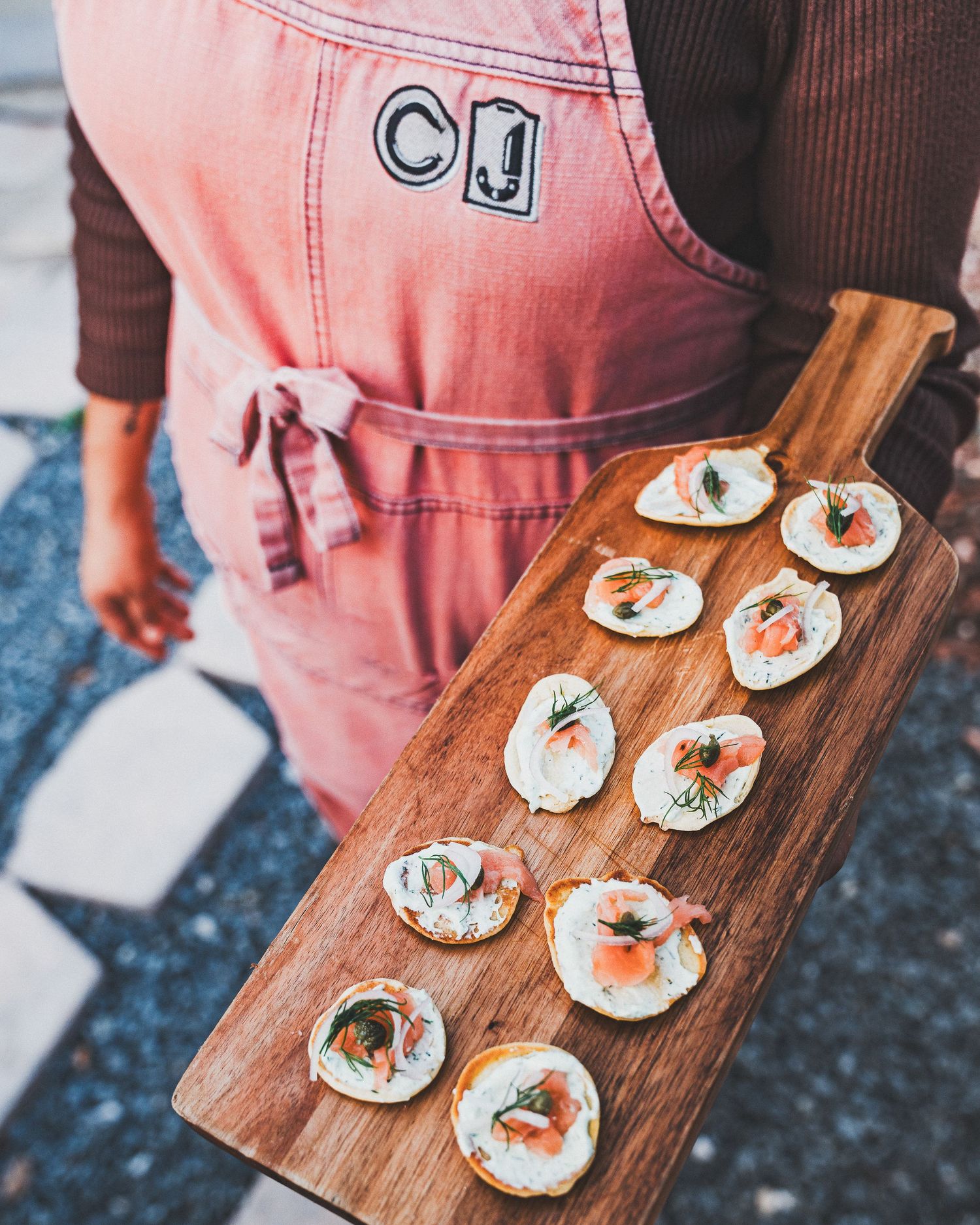 A wooden serving board displays elegant canapés with cream cheese and garnishes held by someone wearing a pink apron.