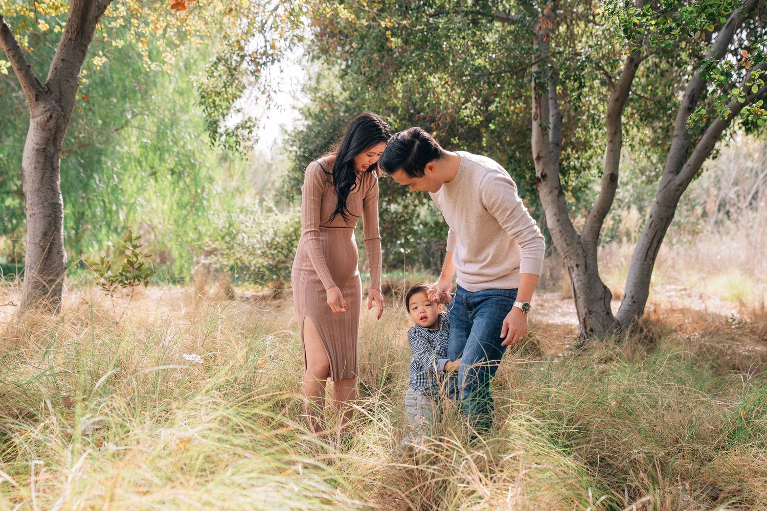 Family walking through a sun-dappled park path with tall grass and trees on a warm summer day.