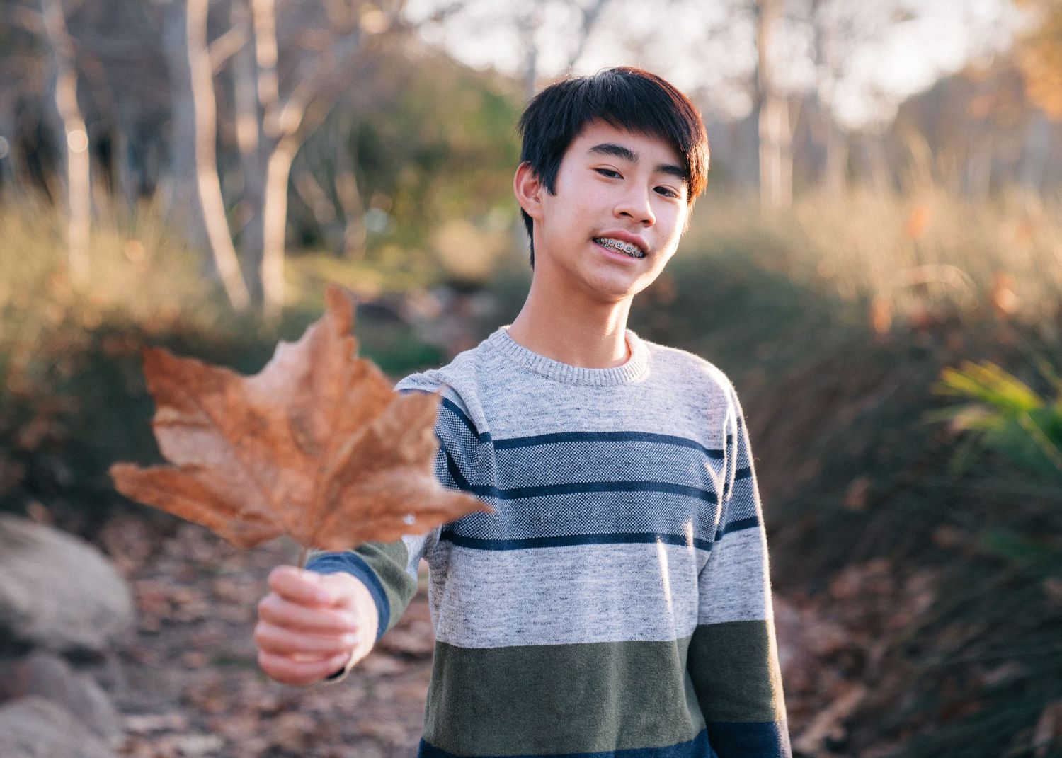 Someone holds up a large autumn leaf while standing outdoors in a striped sweater on a sunny fall day.