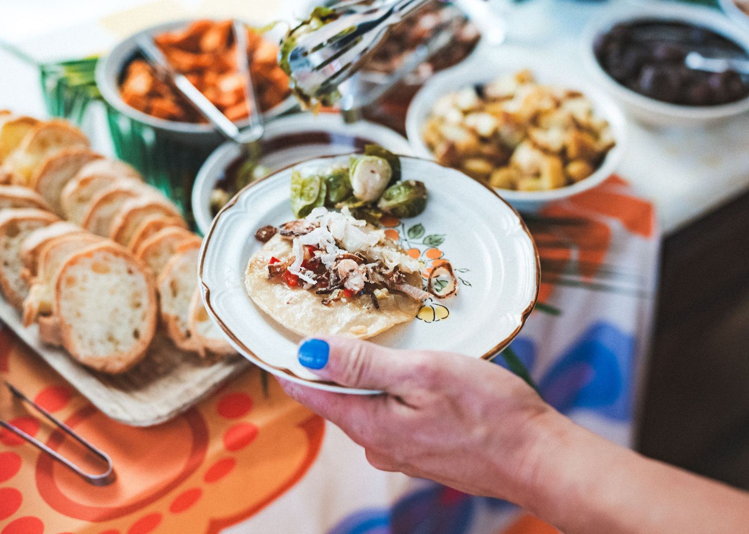 Person holding a white plate of creamy dip with toppings surrounded by bread slices, carrots, and side dishes.