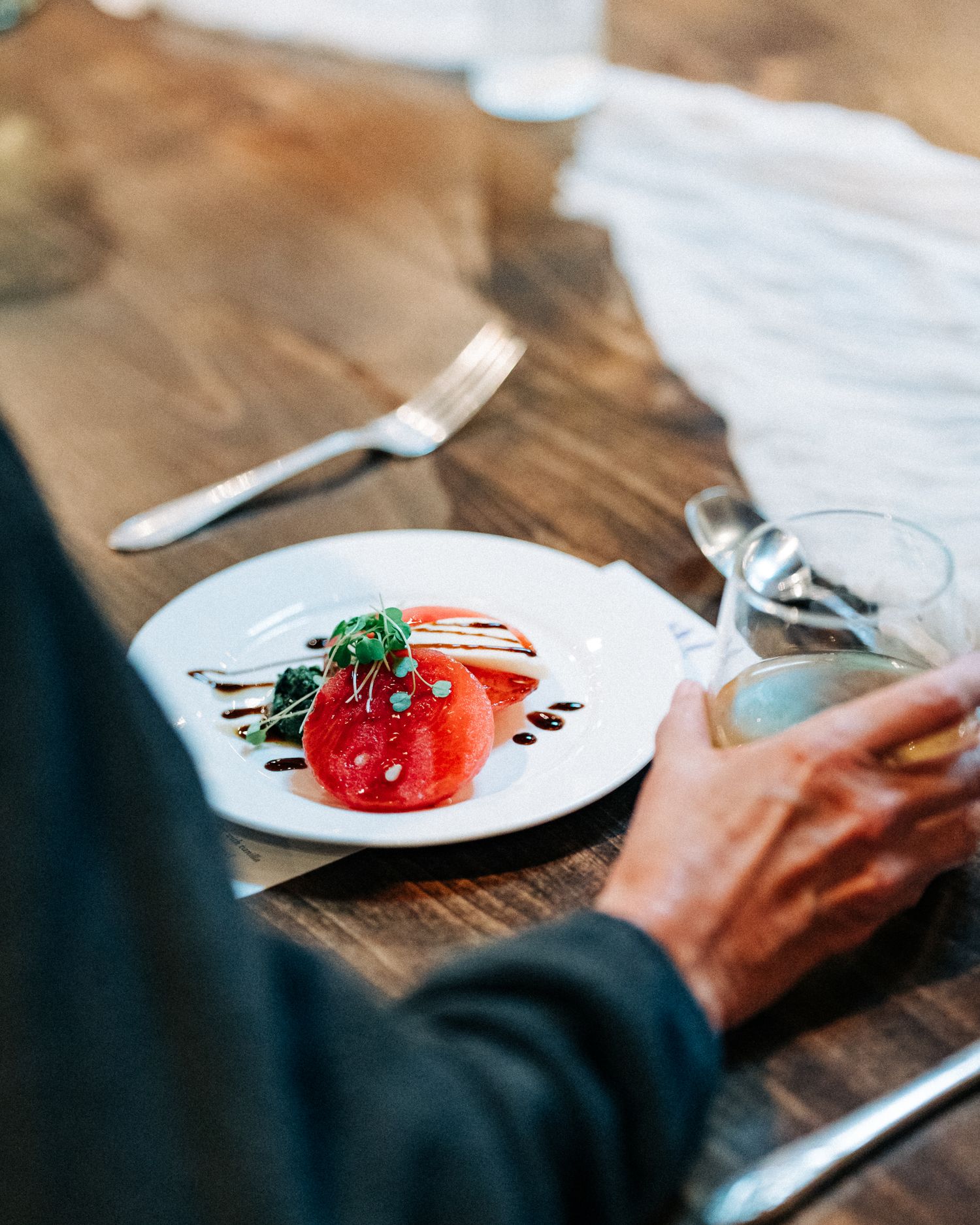 Person dining on a plated appetizer with tomatoes and balsamic drizzle on a rustic wooden table.