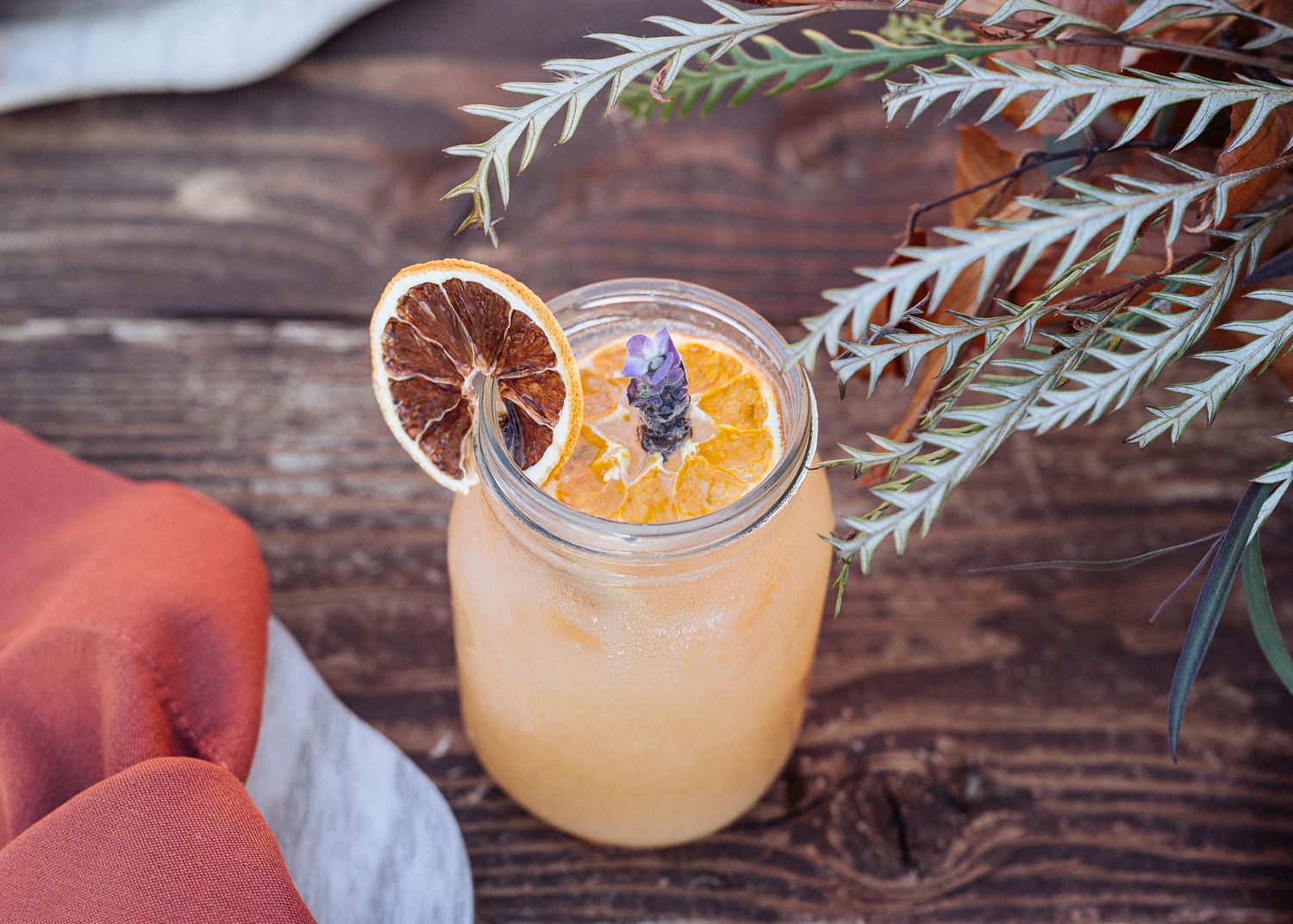 Citrus cocktail garnished with dried orange slice and lavender sprig on wooden surface.