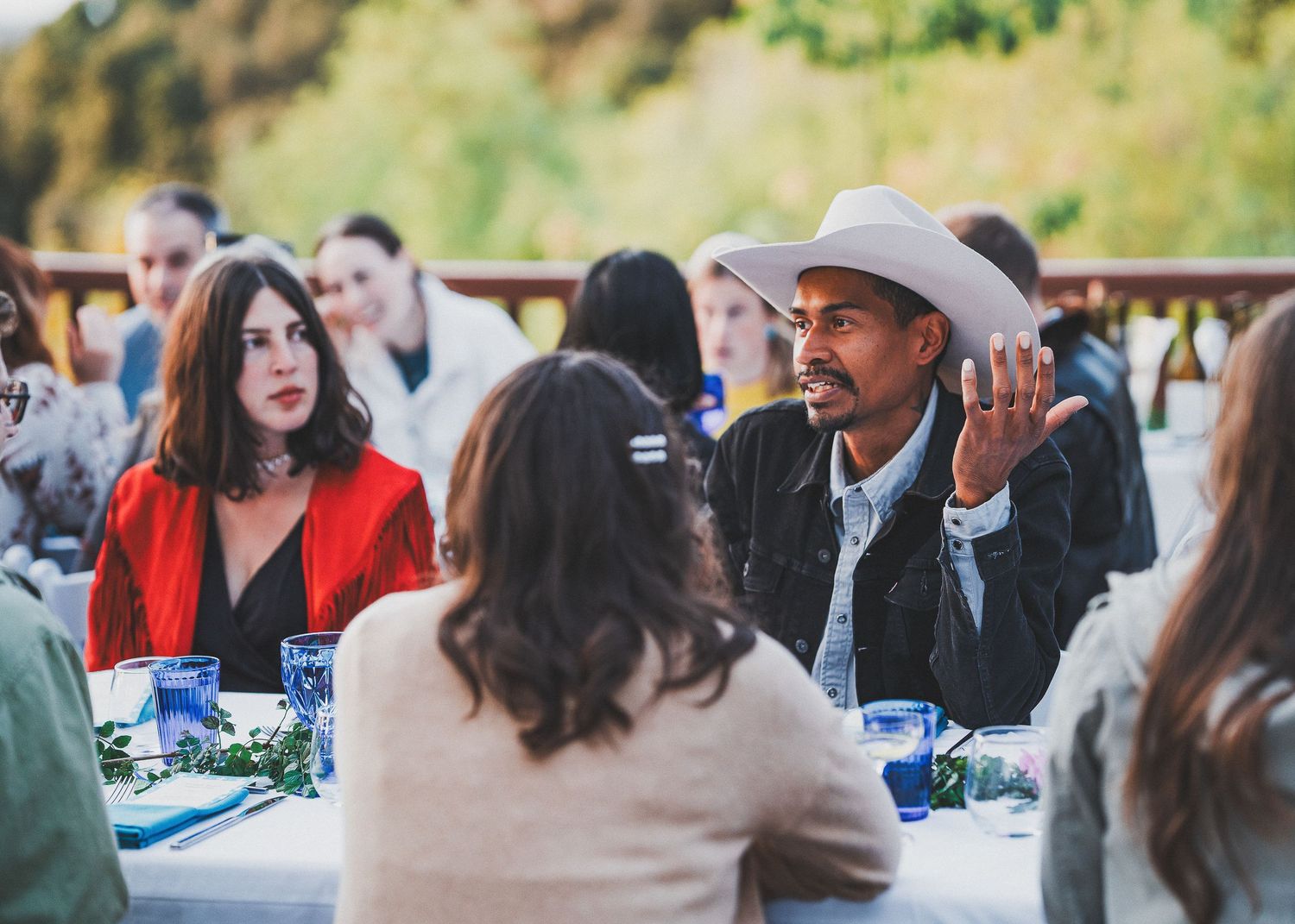 Diverse group of guests engaged in lively conversation at an outdoor dinner party with table settings and natural lighting.